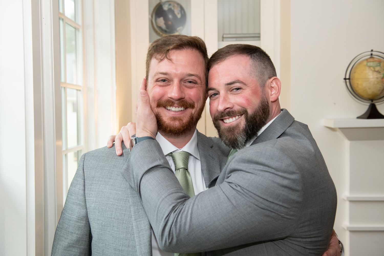 Wedding party members in grey suits share joyful embraces and laughter in a bright indoor setting.