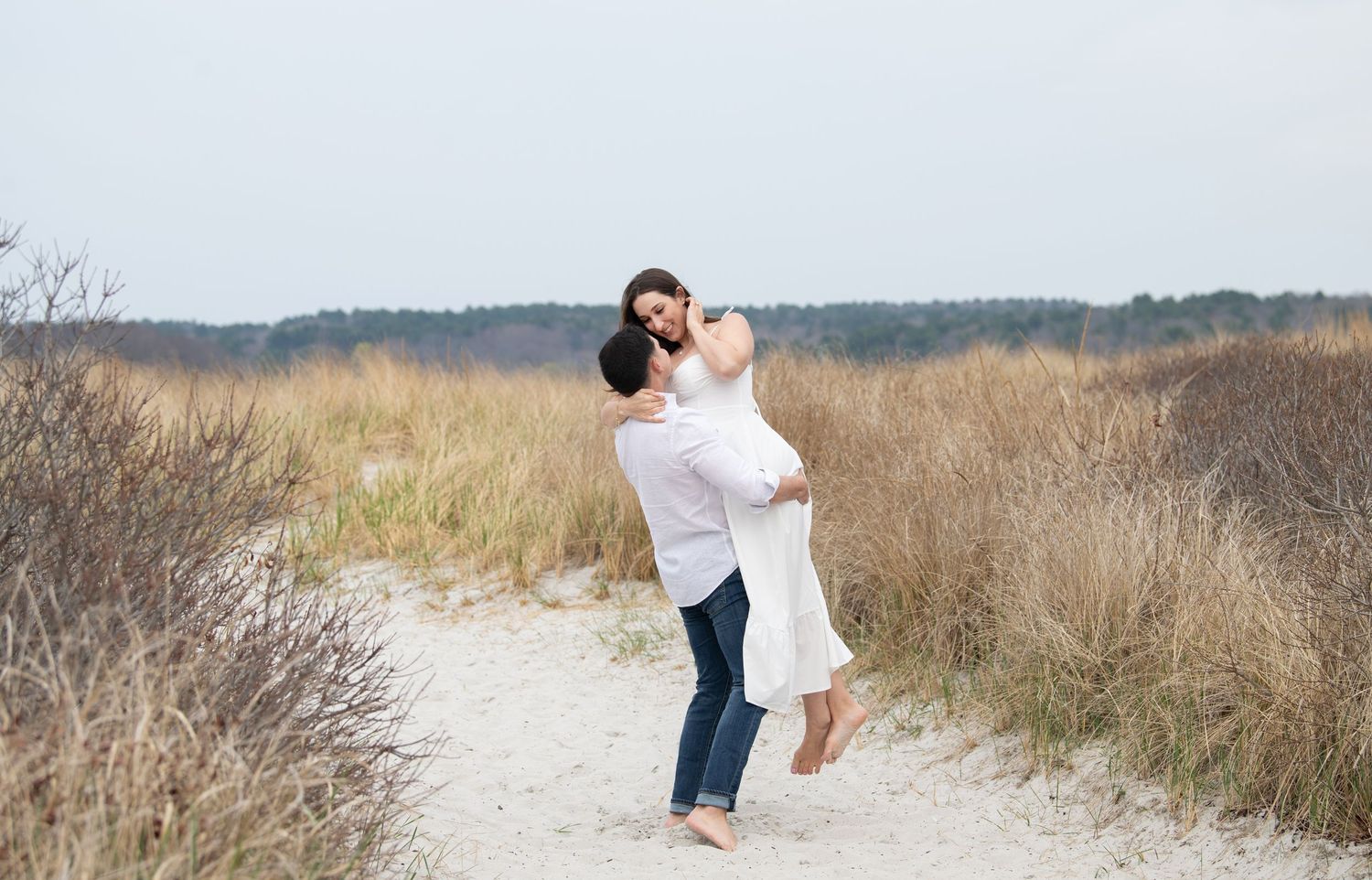 A happy couple embraces and laughs together on a sandy path surrounded by tall dry grass and coastal vegetation.