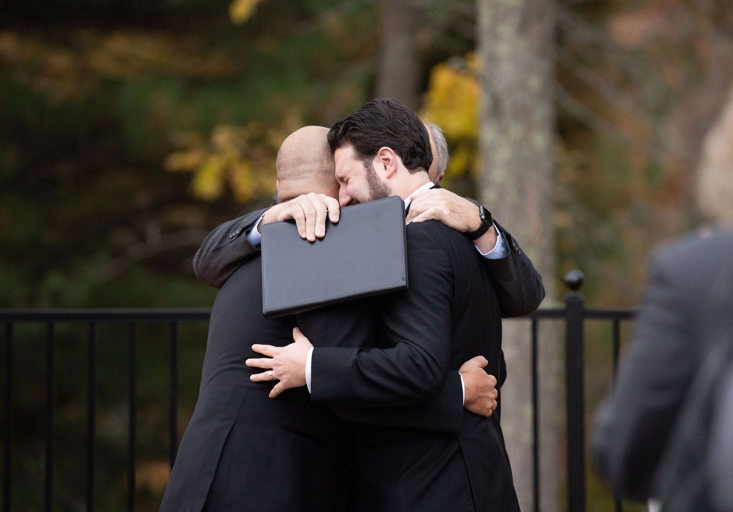 Two people in black formal attire share an emotional embrace outdoors during a special occasion.