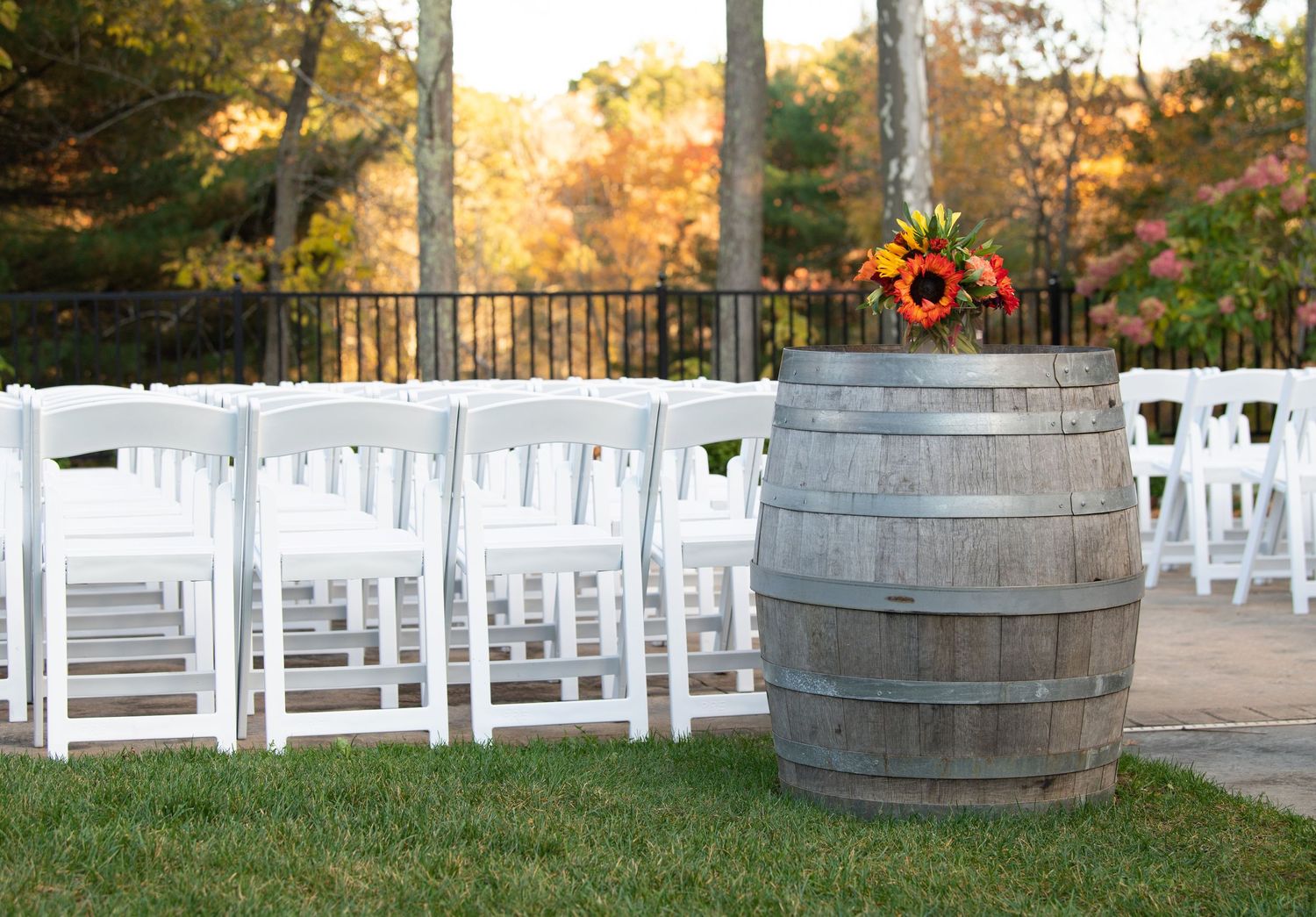 White wooden chairs arranged for an outdoor wedding ceremony with rustic wine barrels and autumn foliage in the background.