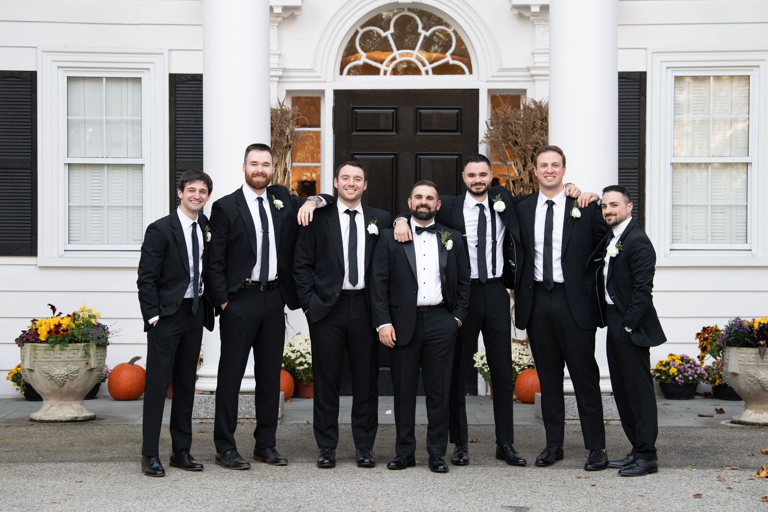 Groomsmen in black suits and ties pose together on white porch steps with autumn pumpkin decorations.