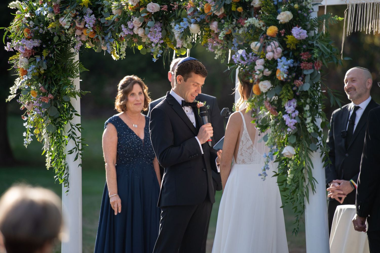 Wedding party stands together under an elegant floral arch decorated with purple and white flowers during outdoor ceremony.