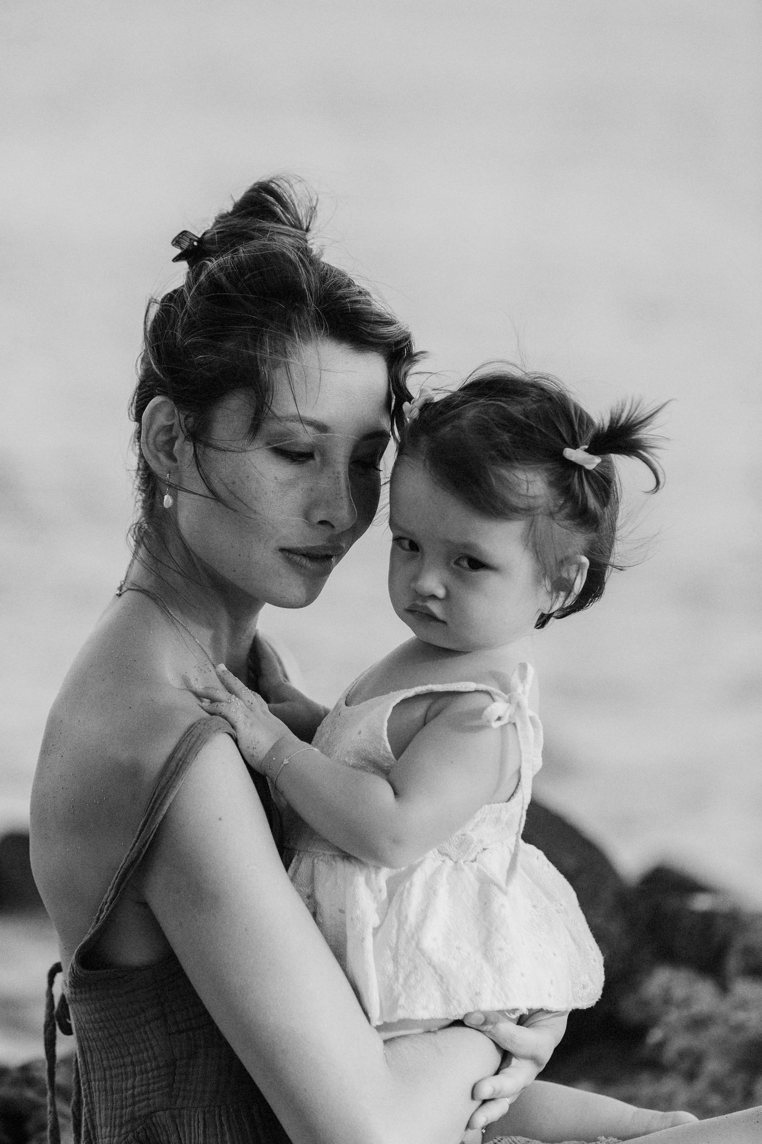 A black and white portrait of a mother tenderly holding her baby daughter in a white dress outdoors.