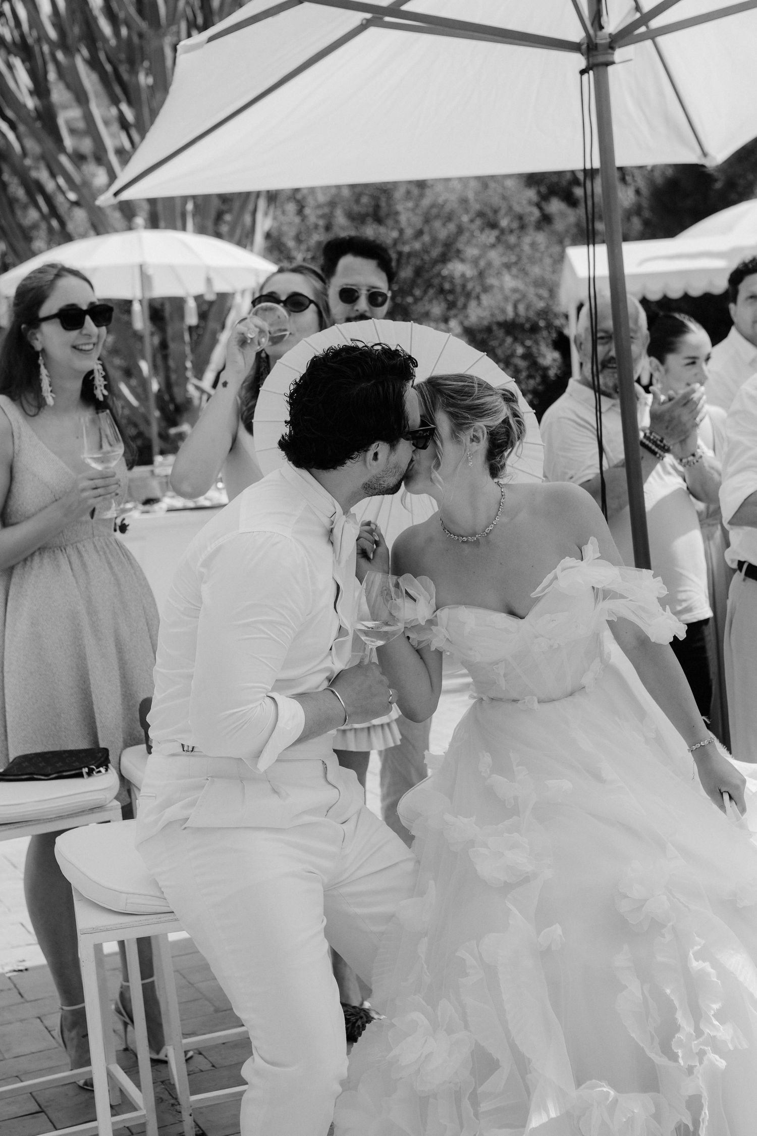 A group of people in white attire gather under umbrellas at an outdoor event on a sunny day.