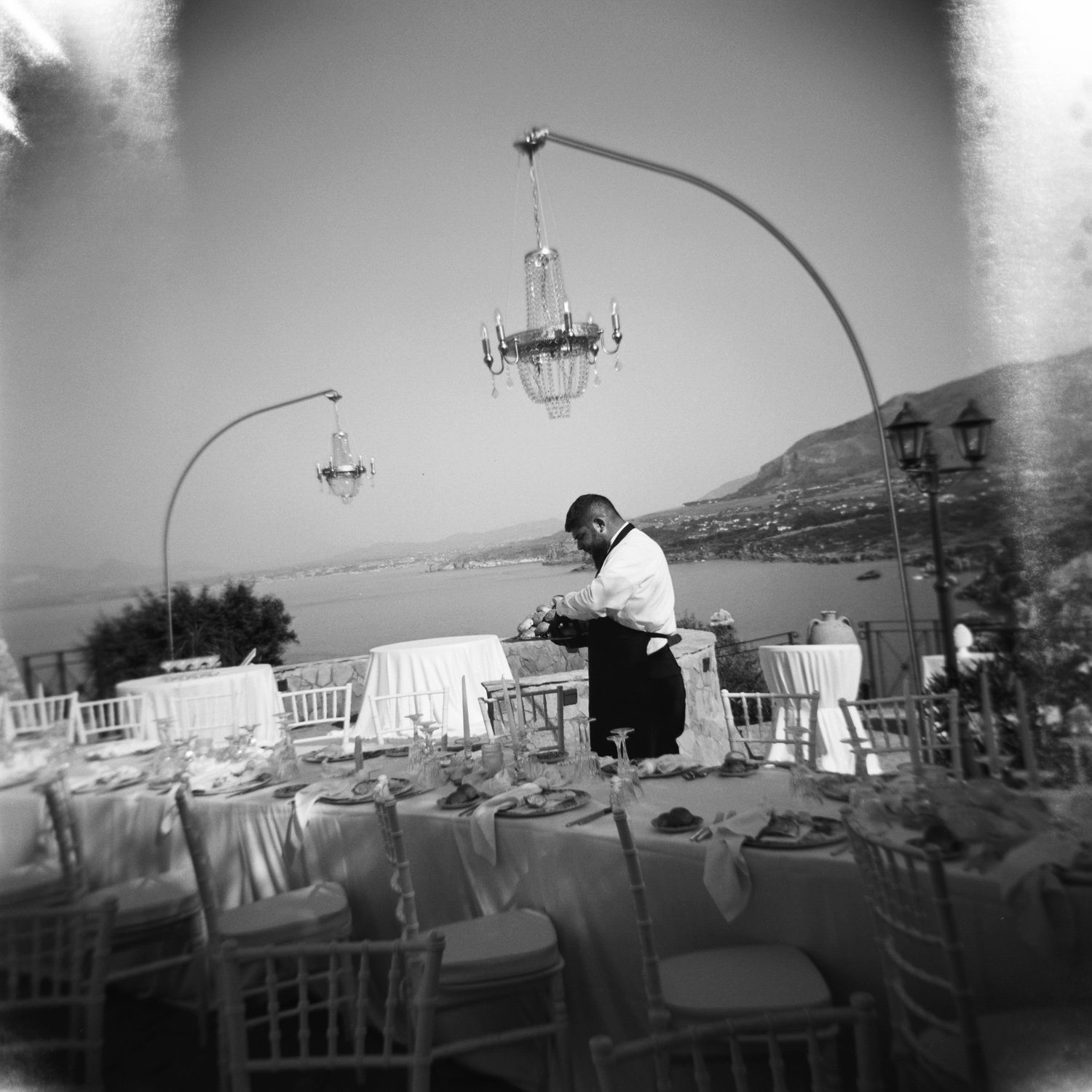 Black and white panoramic sequence showing an outdoor restaurant terrace with white tables overlooking a coastal landscape.