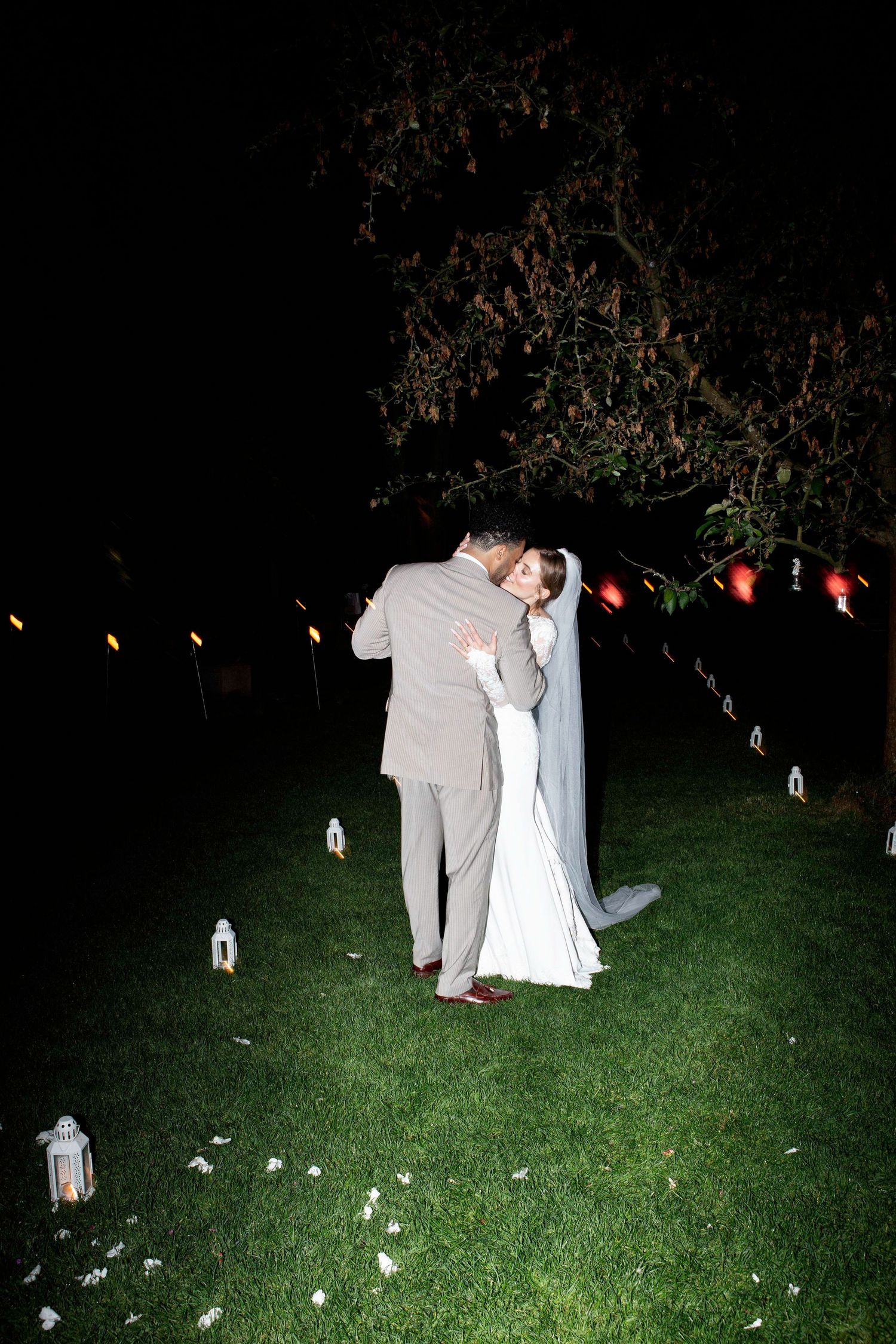 A romantic couple in formal attire shares a tender moment on a dark lawn decorated with glowing lanterns.