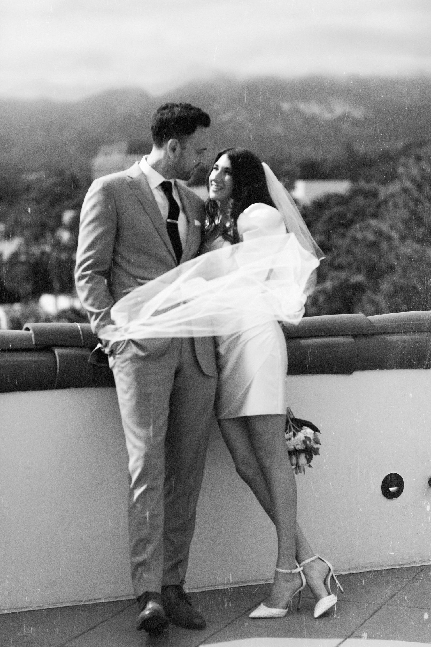 Black and white photo of a bride and groom sharing a romantic moment on a rooftop, her veil flowing in the breeze.