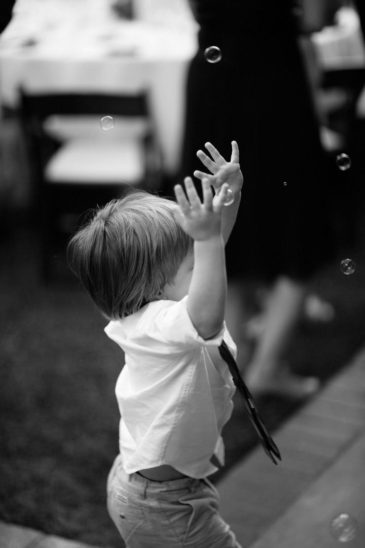 Black and white photo of a toddler reaching up to catch soap bubbles, wearing a white shirt.