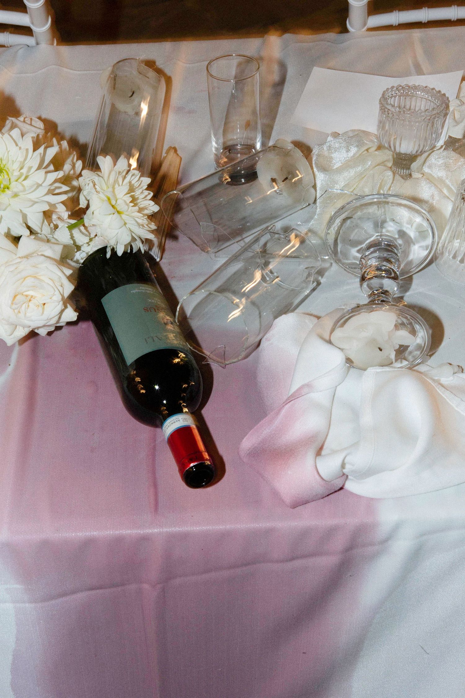 Tipped over wine glasses and white flowers on a pink tablecloth create an artistic still life composition.