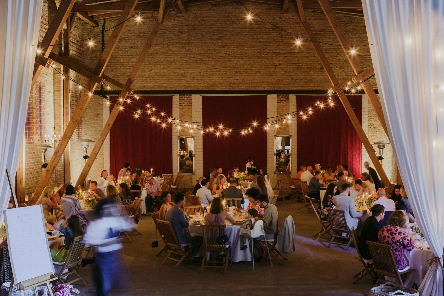 Rustic barn wedding reception venue decorated with string lights and white drapes showing guests seated at round tables.