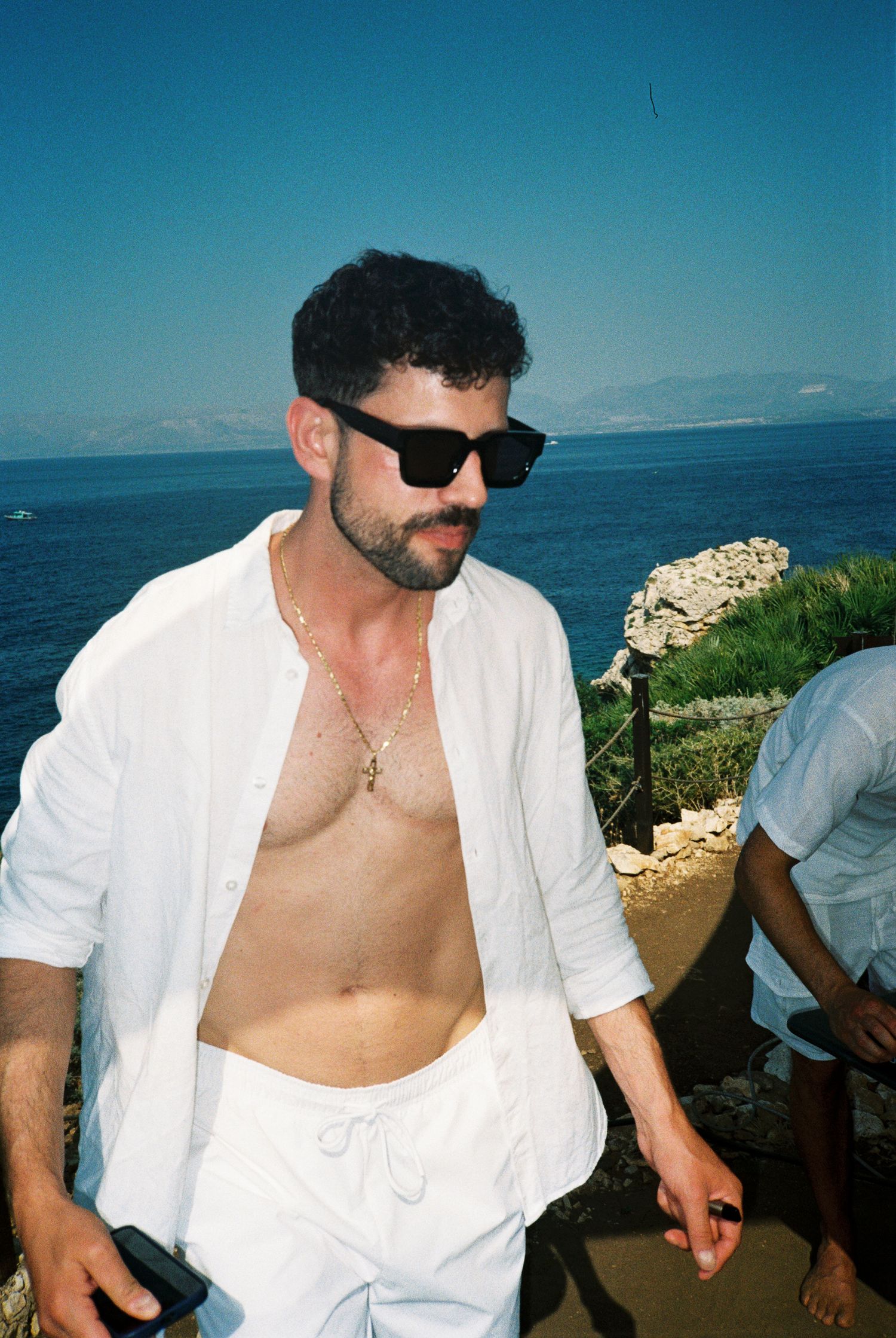 Person in white open shirt poses against scenic ocean backdrop on sunny day.