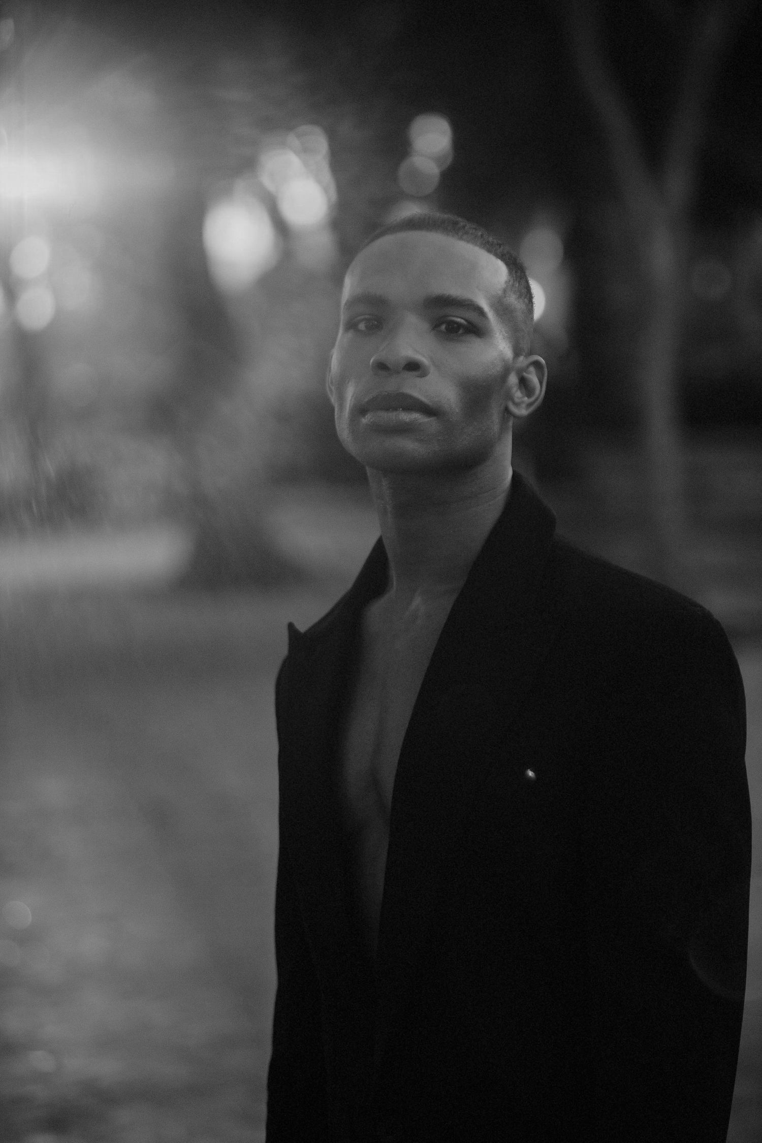 Black and white portrait of a young man in an open blazer outdoors at night with bokeh lights.