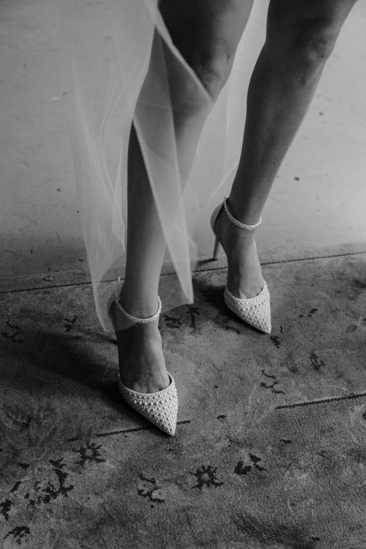 Black and white photo of a bride wearing embellished pearl heels with ankle straps and a flowing tulle wedding dress.