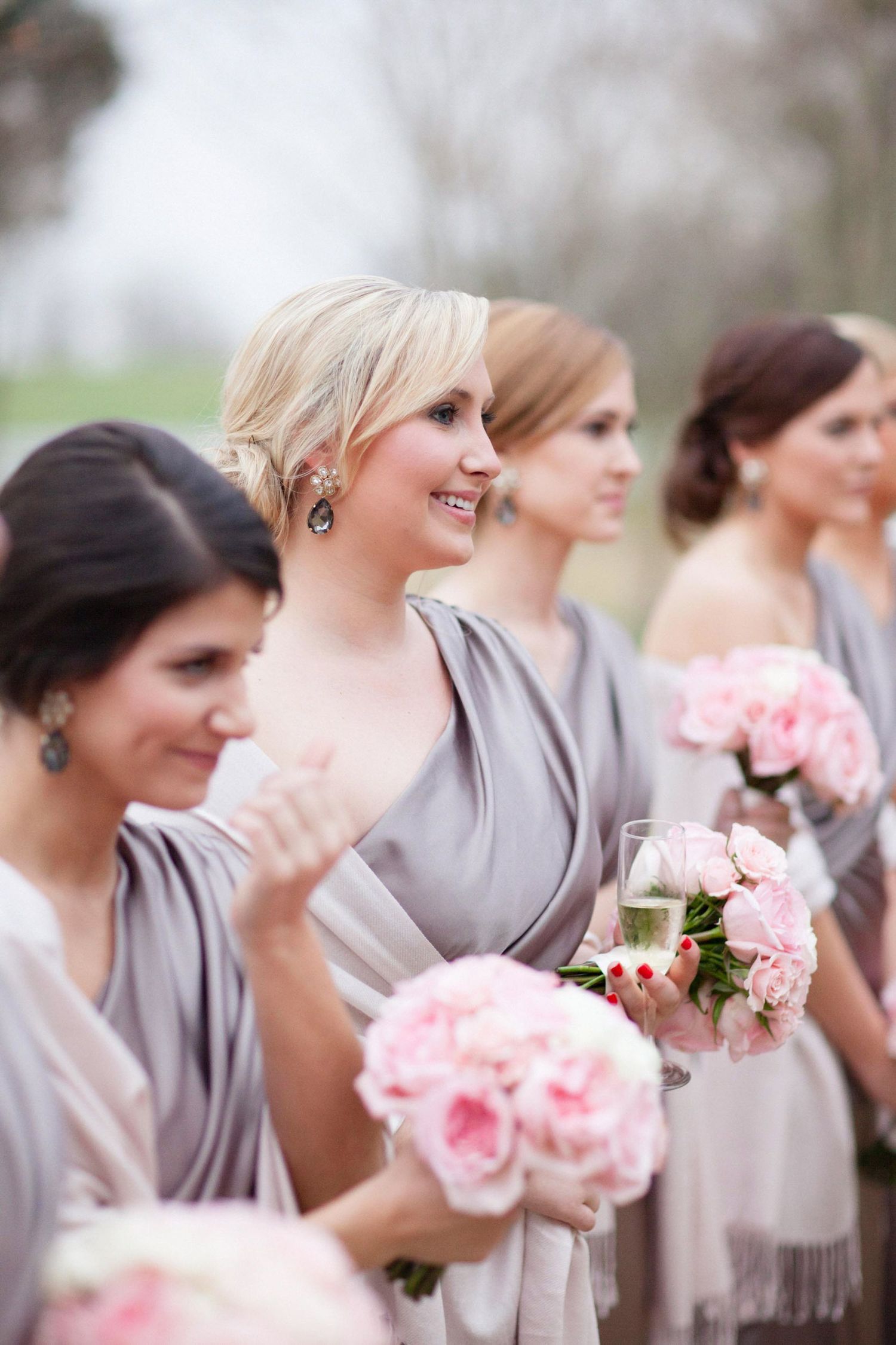 Row of bridesmaids in gray dresses holding pink rose bouquets stand together at an outdoor wedding ceremony.