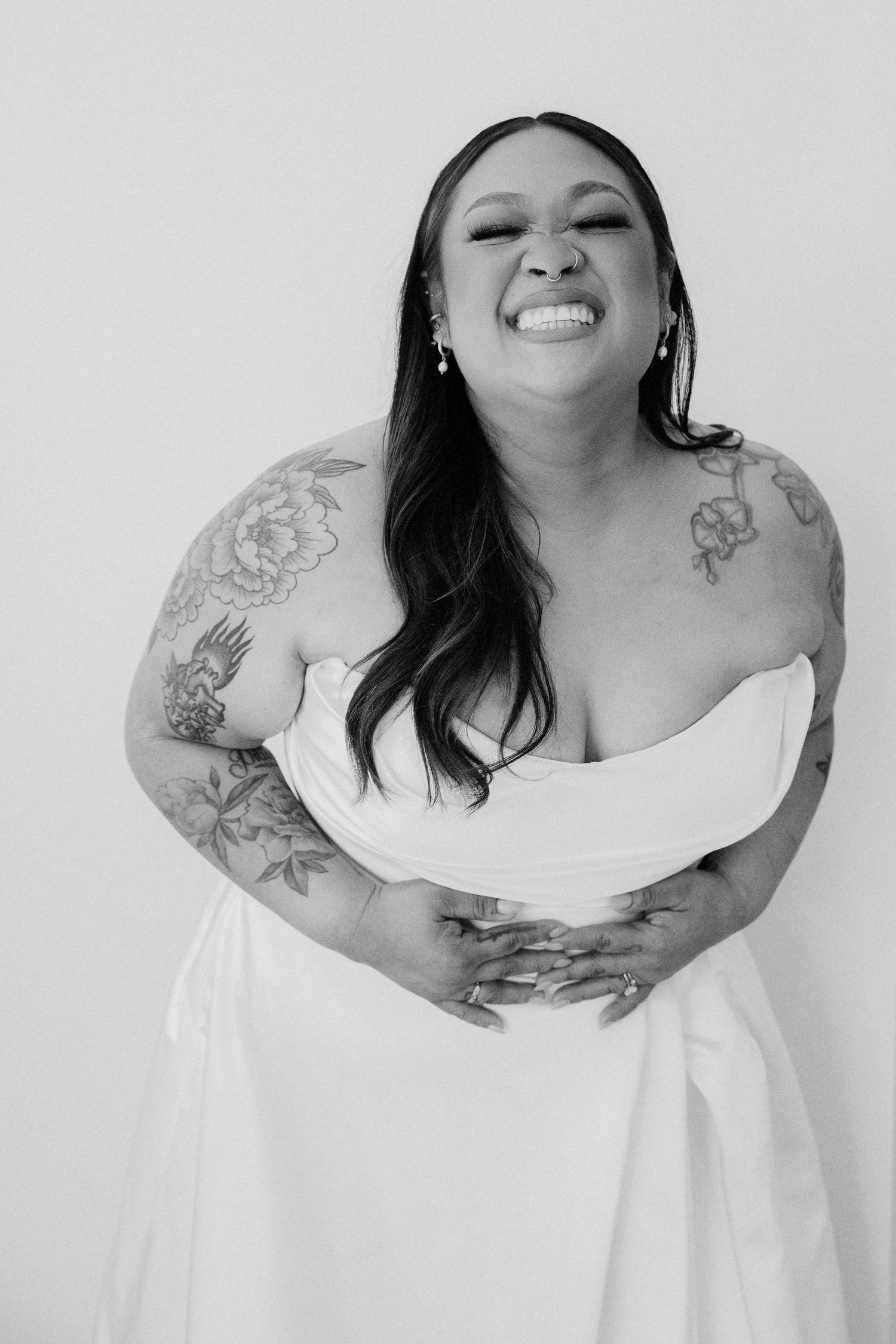 Black and white photo of a laughing bride in strapless white gown with floral tattoos on her arms.