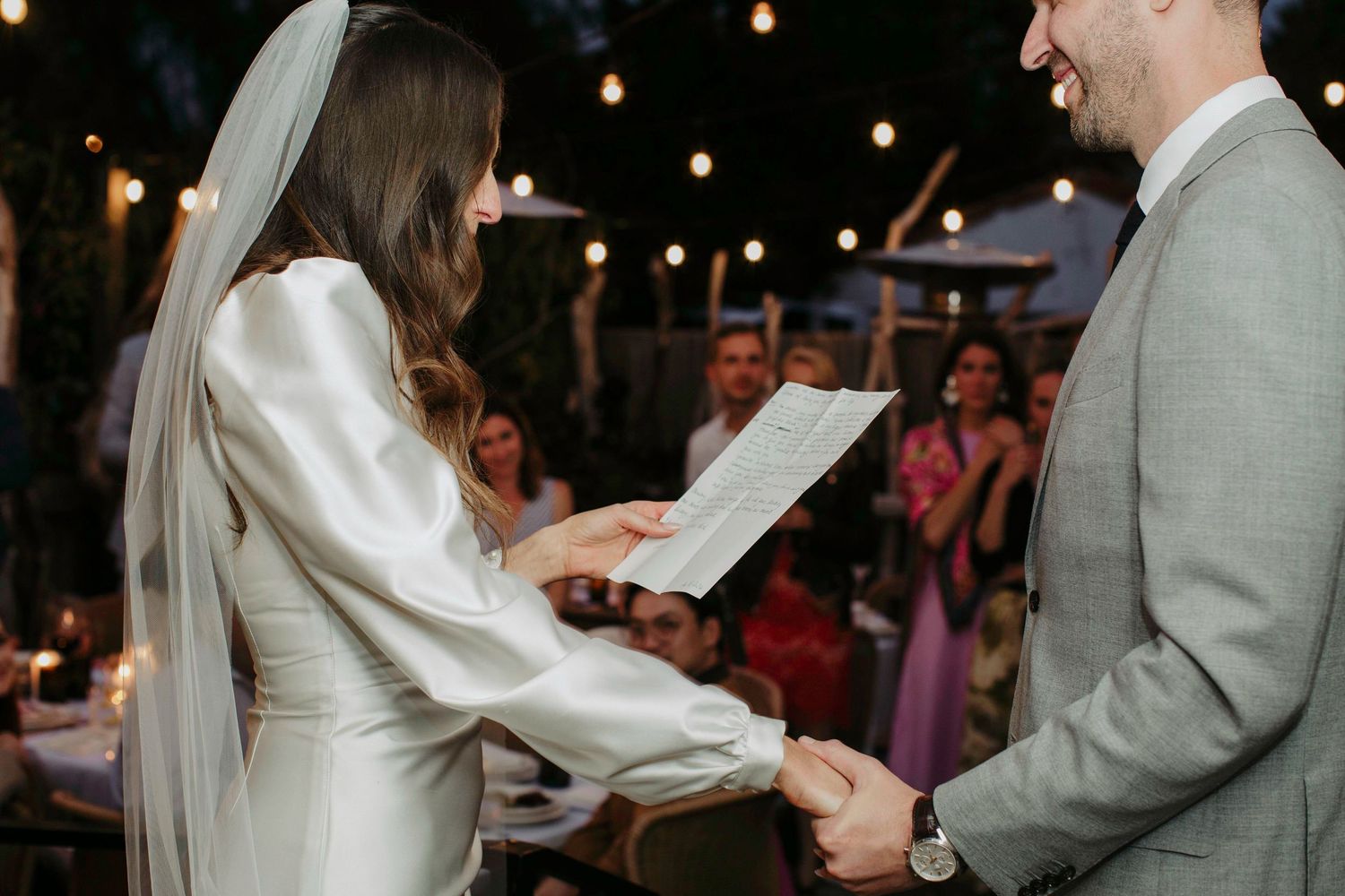 Bride reading wedding vows to groom outdoors at romantic evening ceremony with string lights.