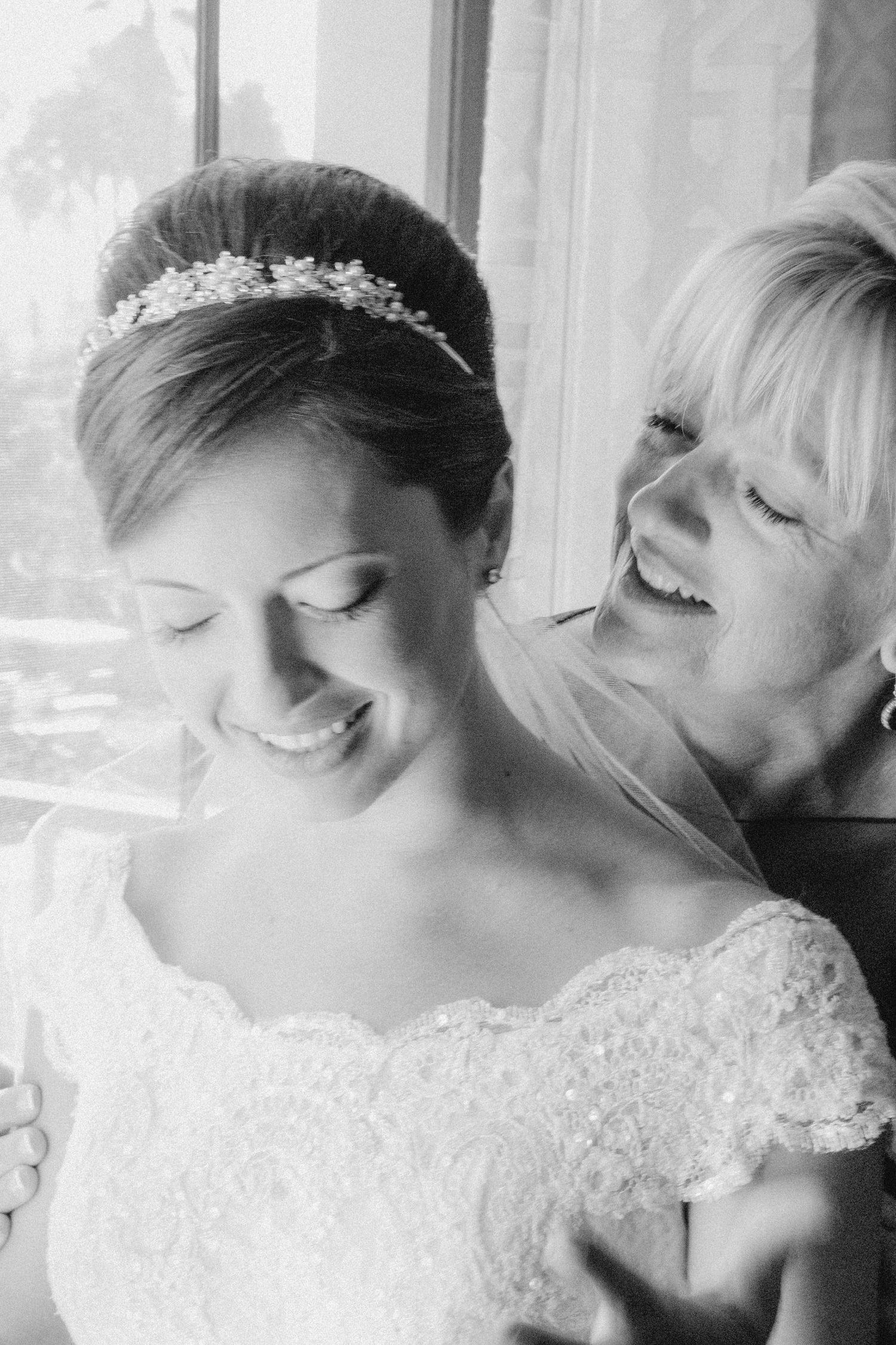 Black and white photo of a smiling bride in lace gown and tiara being helped by a laughing older woman near a window.