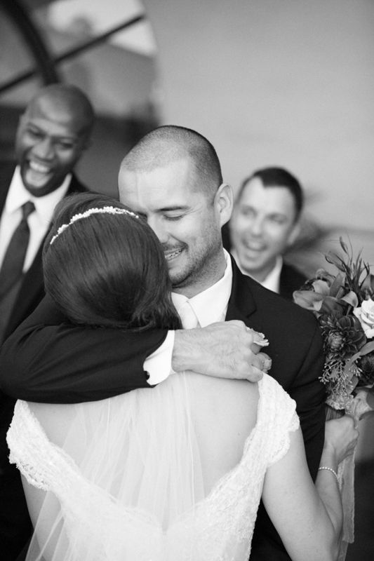 Black and white photo of a groom embracing his bride at a wedding ceremony with smiling groomsmen in the background.