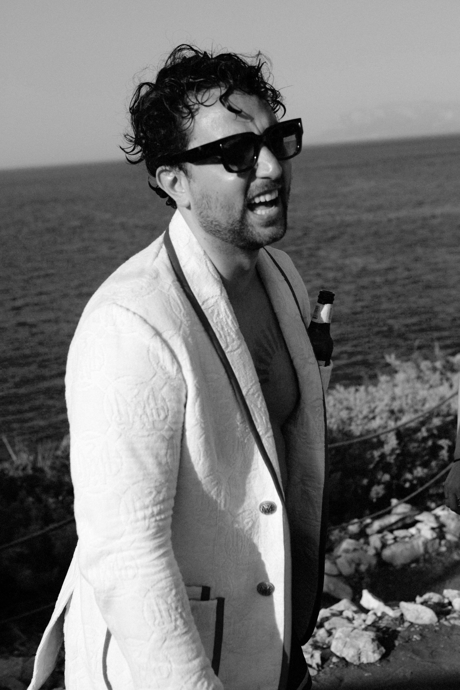 A person in a white jacket and sunglasses stands against a rocky coastal backdrop on a sunny day.