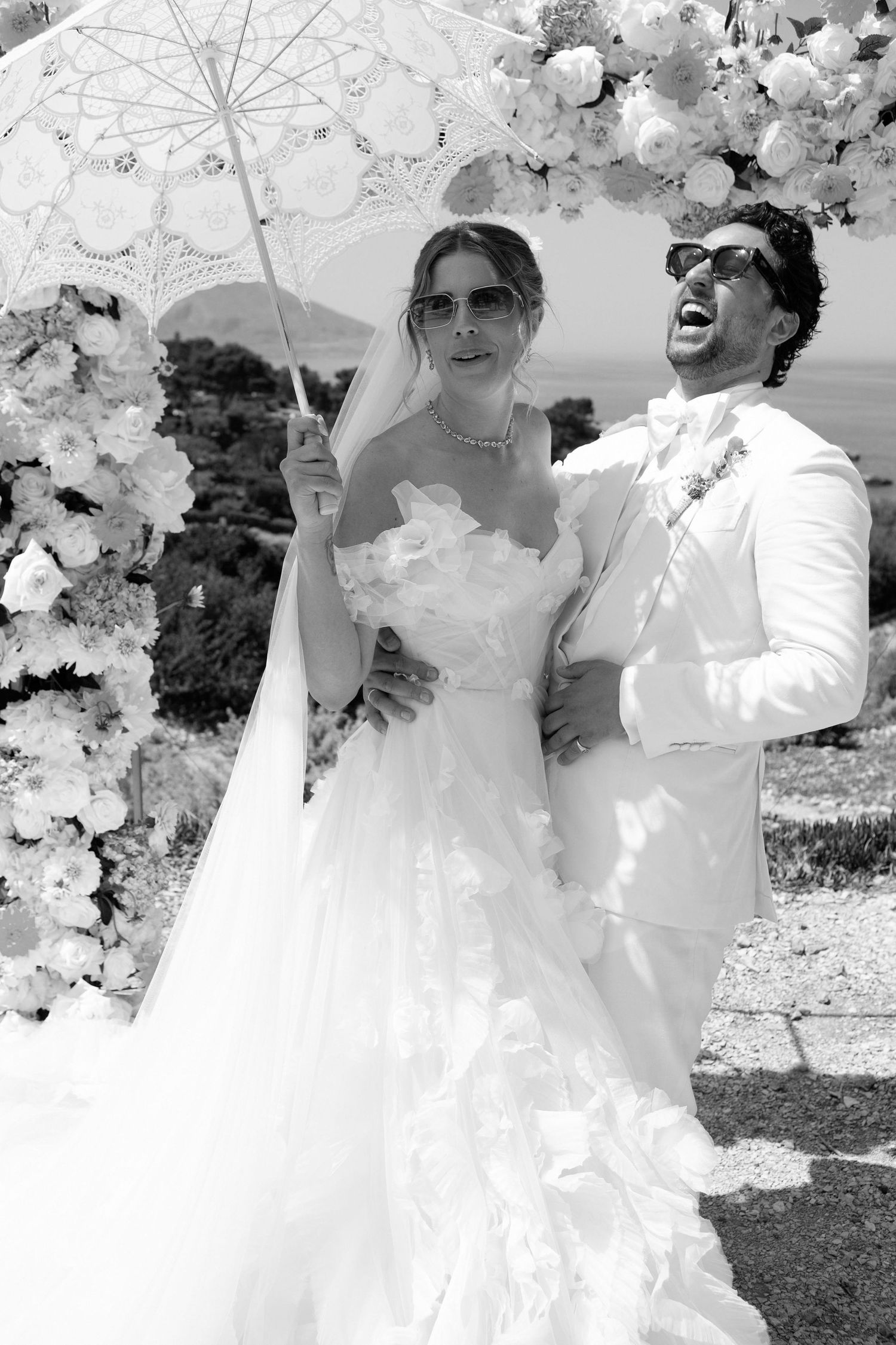 A romantic wedding couple embraces outdoors under white cherry blossoms on a sunny spring day.