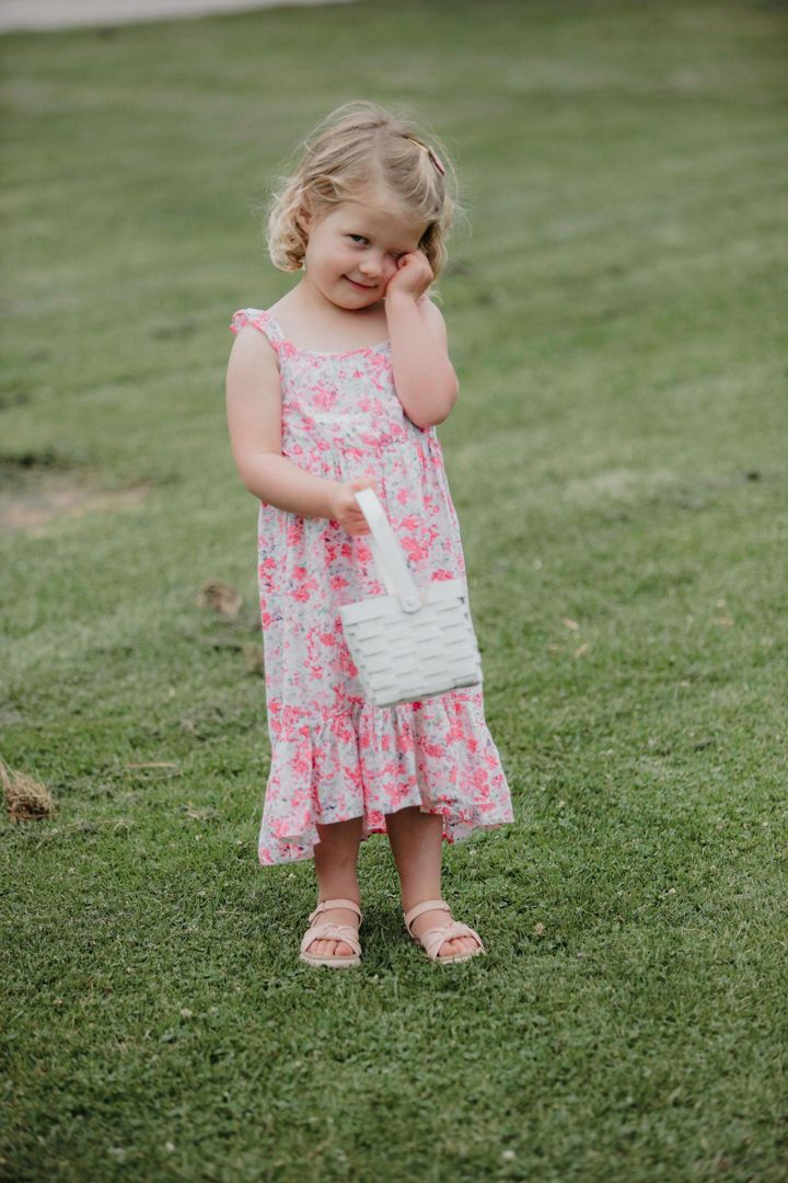 A small child in a pink floral sundress and sandals stands on grass while holding a white purse.