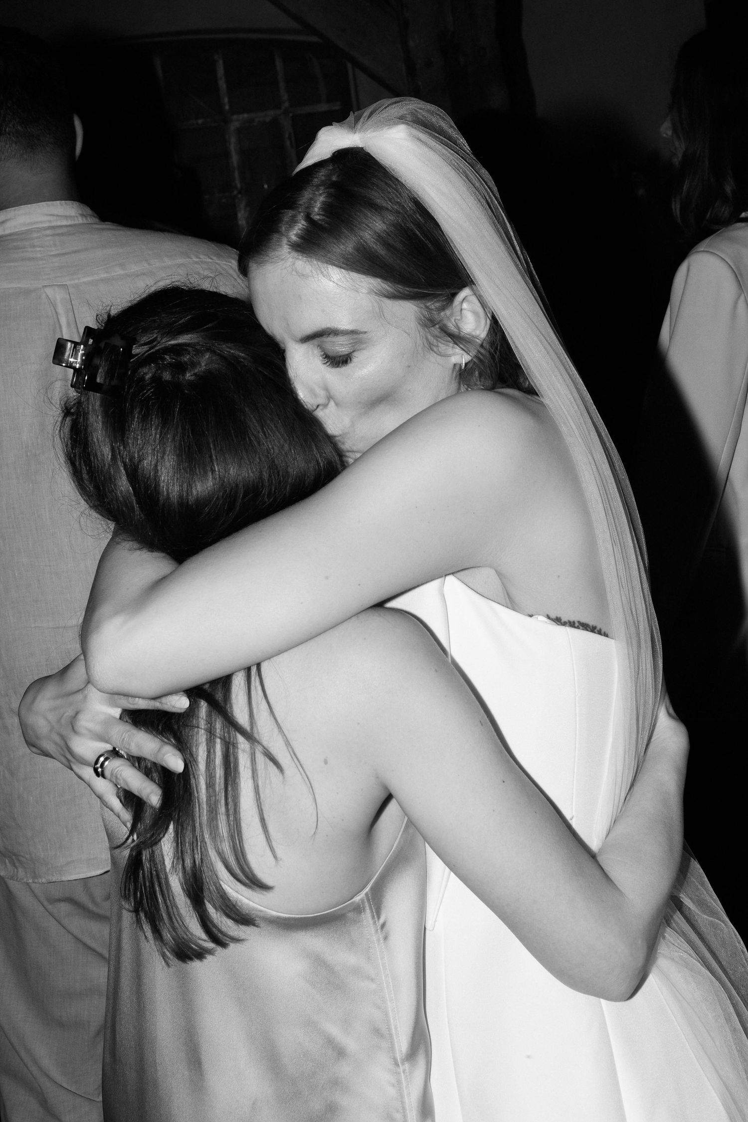 Black and white photo of a bride in white dress and veil sharing a warm embrace with a guest at her wedding reception.