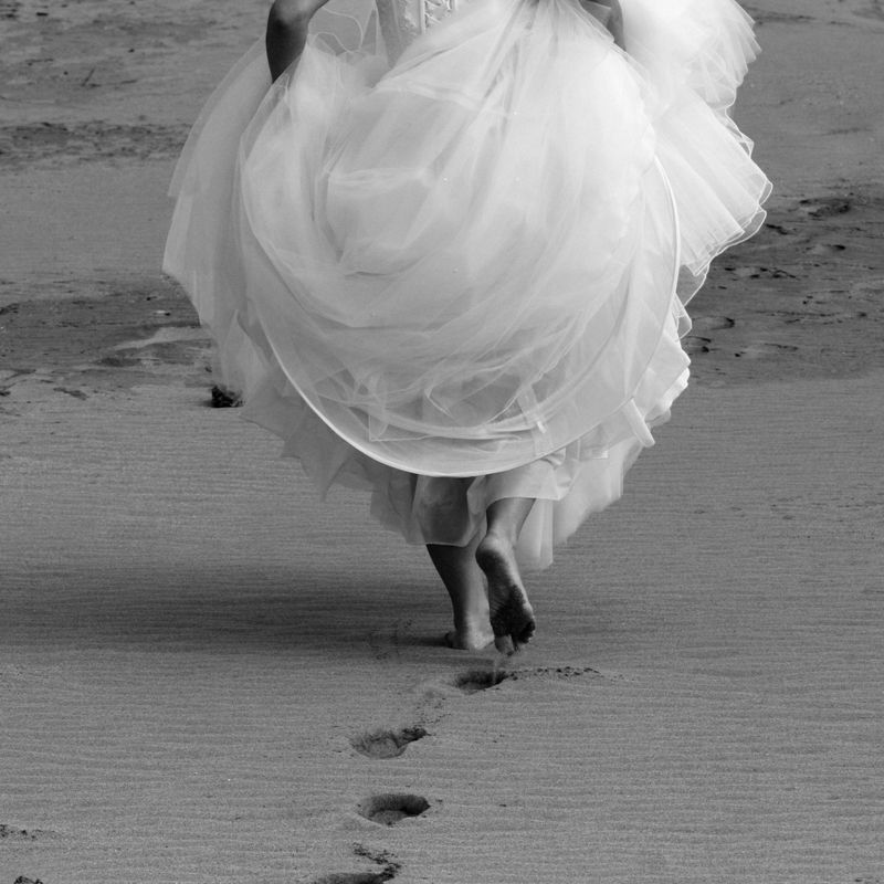 Black and white image of a flowing white dress and feet walking on a sandy beach, creating an ethereal effect.