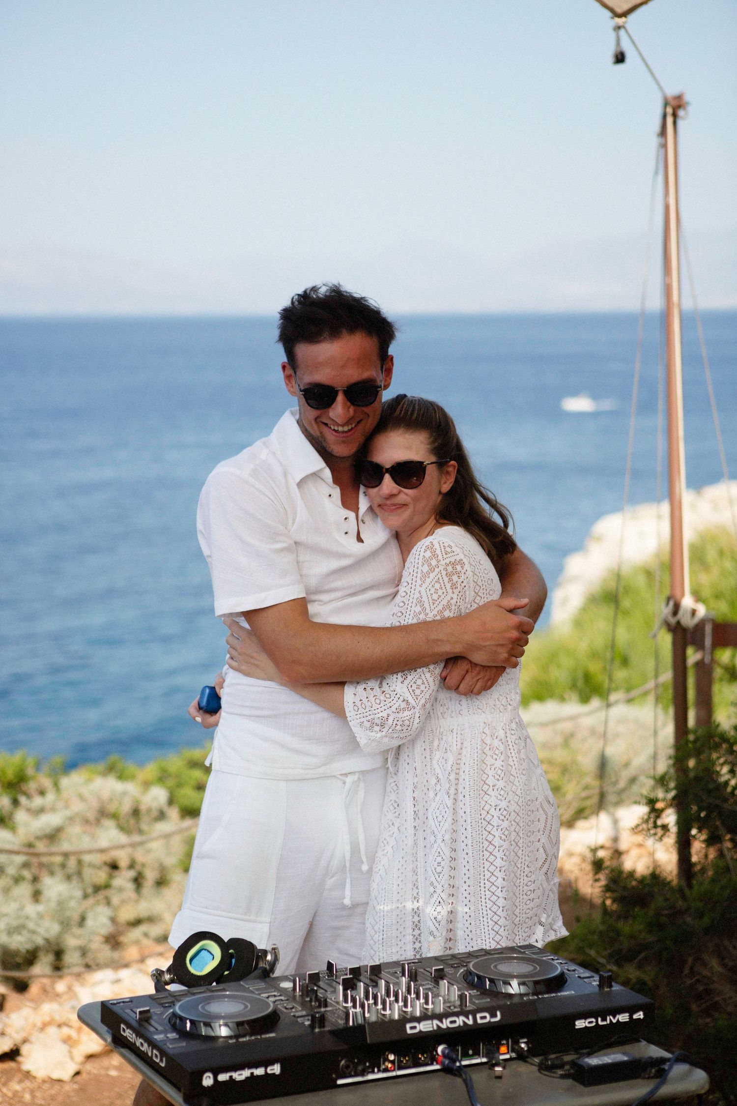 DJ couple in white outfits embrace while performing at an outdoor venue overlooking the ocean.