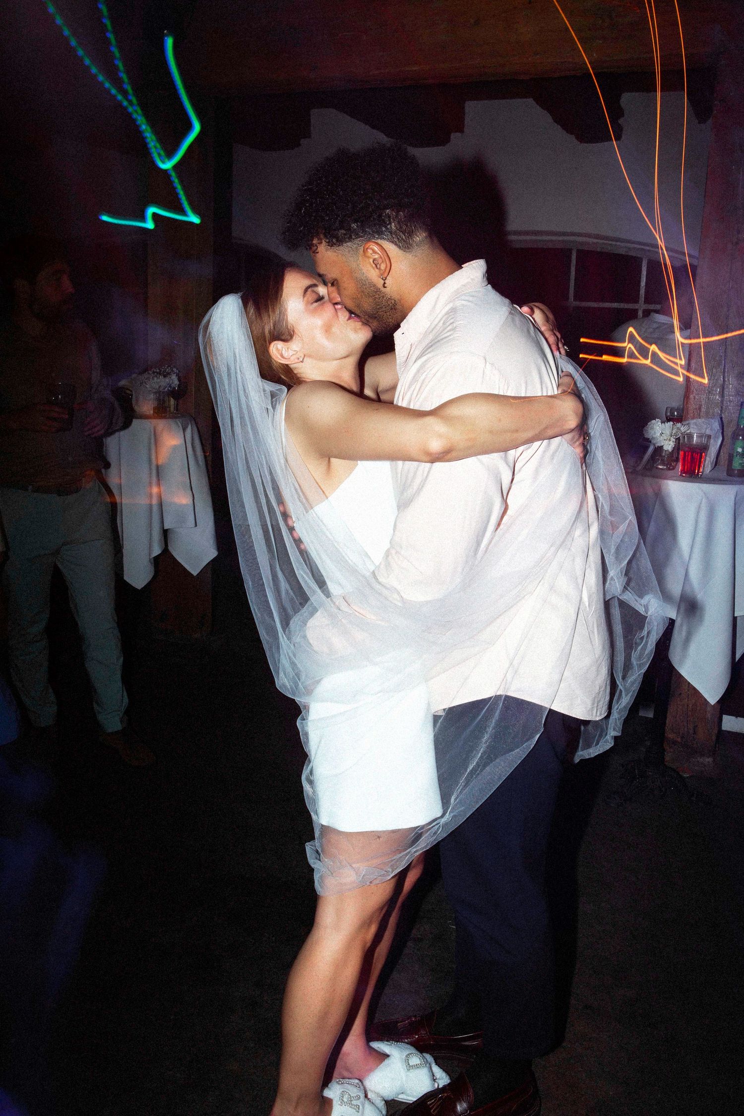 A couple in wedding attire share an intimate dance together at a dimly lit reception venue with colorful lights.