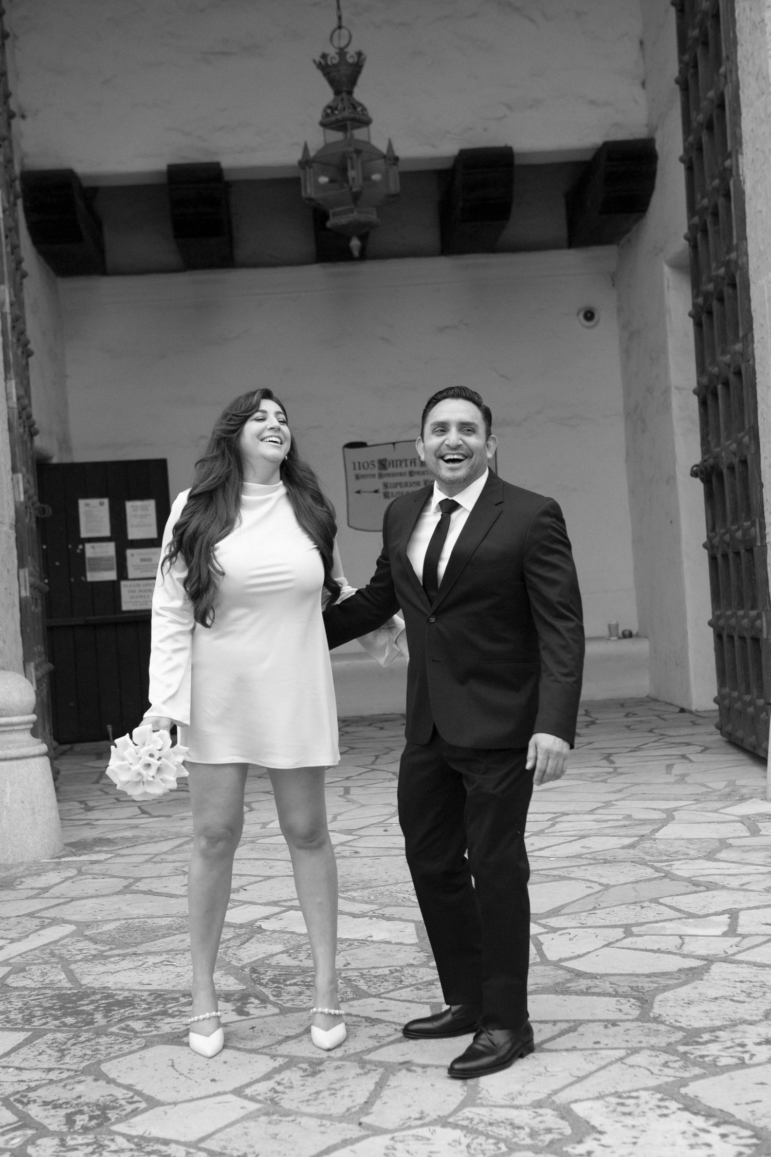 A black and white photo of a couple in formal attire standing together on a patio with stone flooring.
