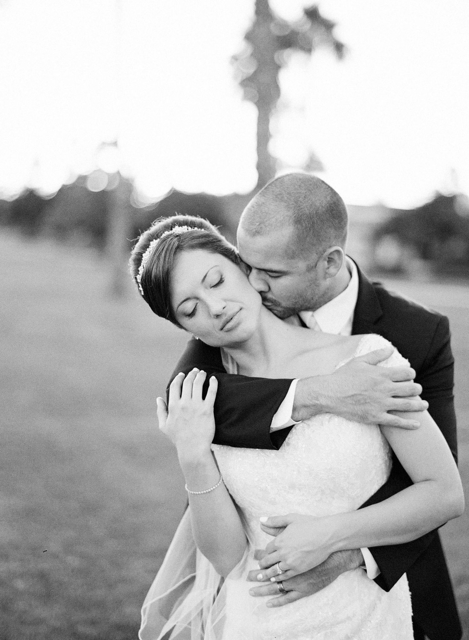 Black and white wedding photo of groom kissing bride on cheek while embracing outdoors near palm tree.