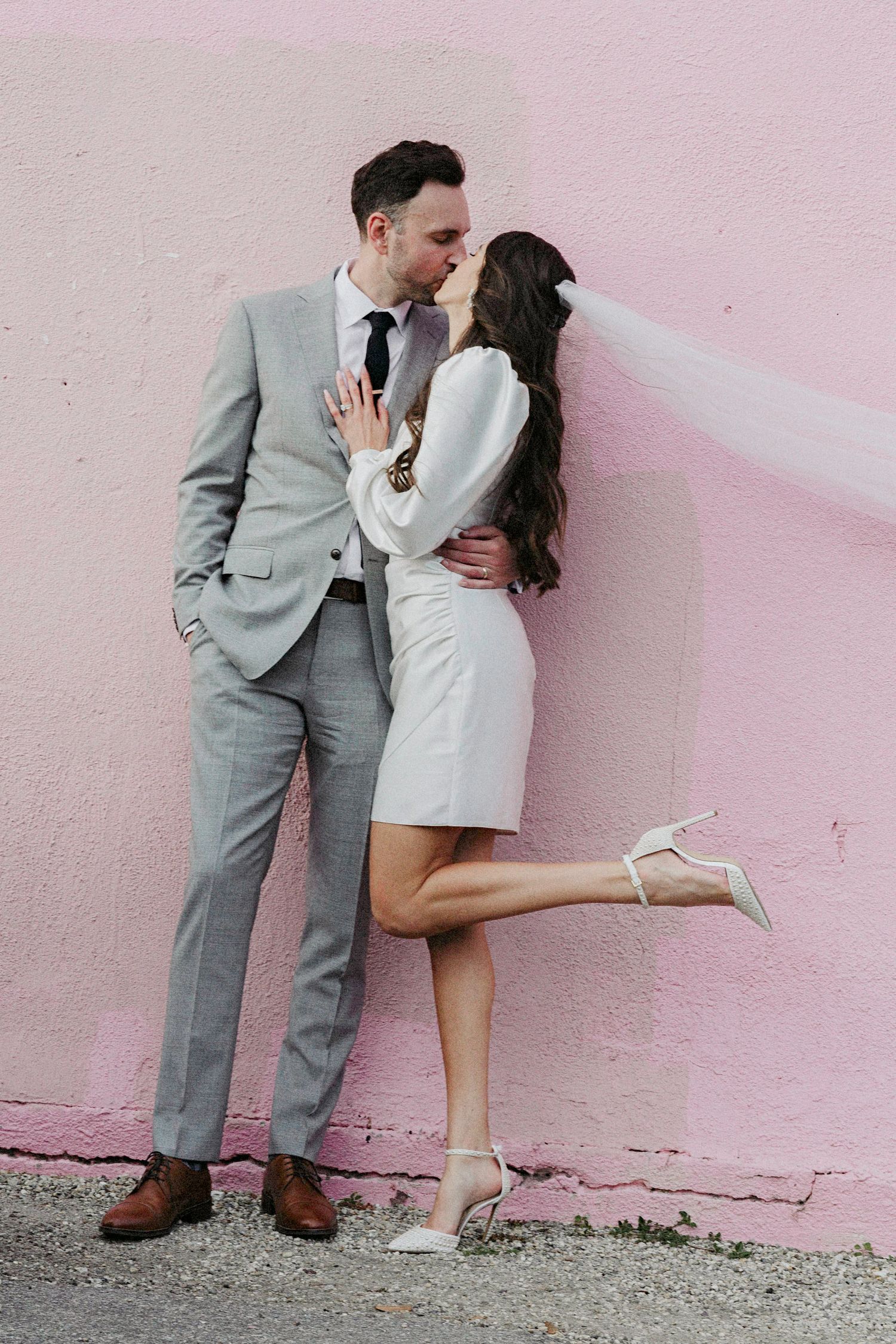 A romantic couple in gray suit and white dress share intimate moments against a pink wall in a sequence of photos.