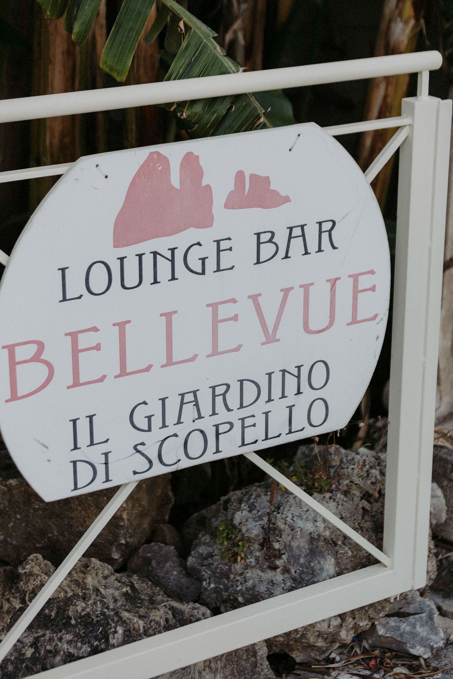 White directional signs with Italian text 'Il Giardino di Scopello' and 'Bellevue' mounted on a stone wall.