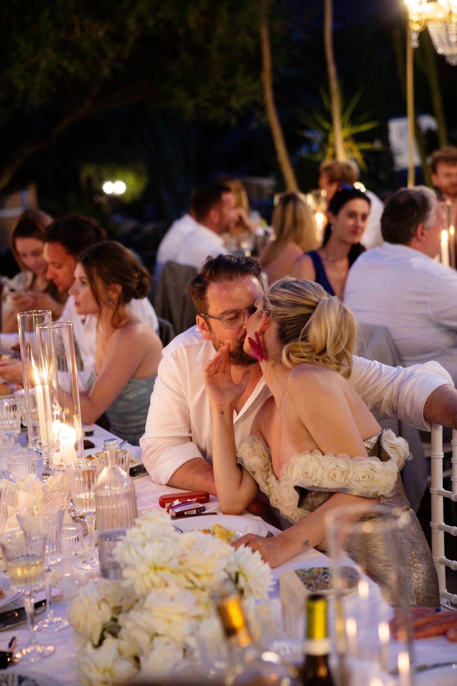 Wedding reception guests seated at a long table with candlelight and white floral centerpieces during evening dinner.