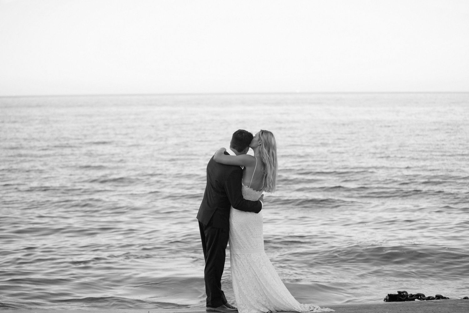 Black and white photo of a bride and groom embracing by the ocean on their wedding day.