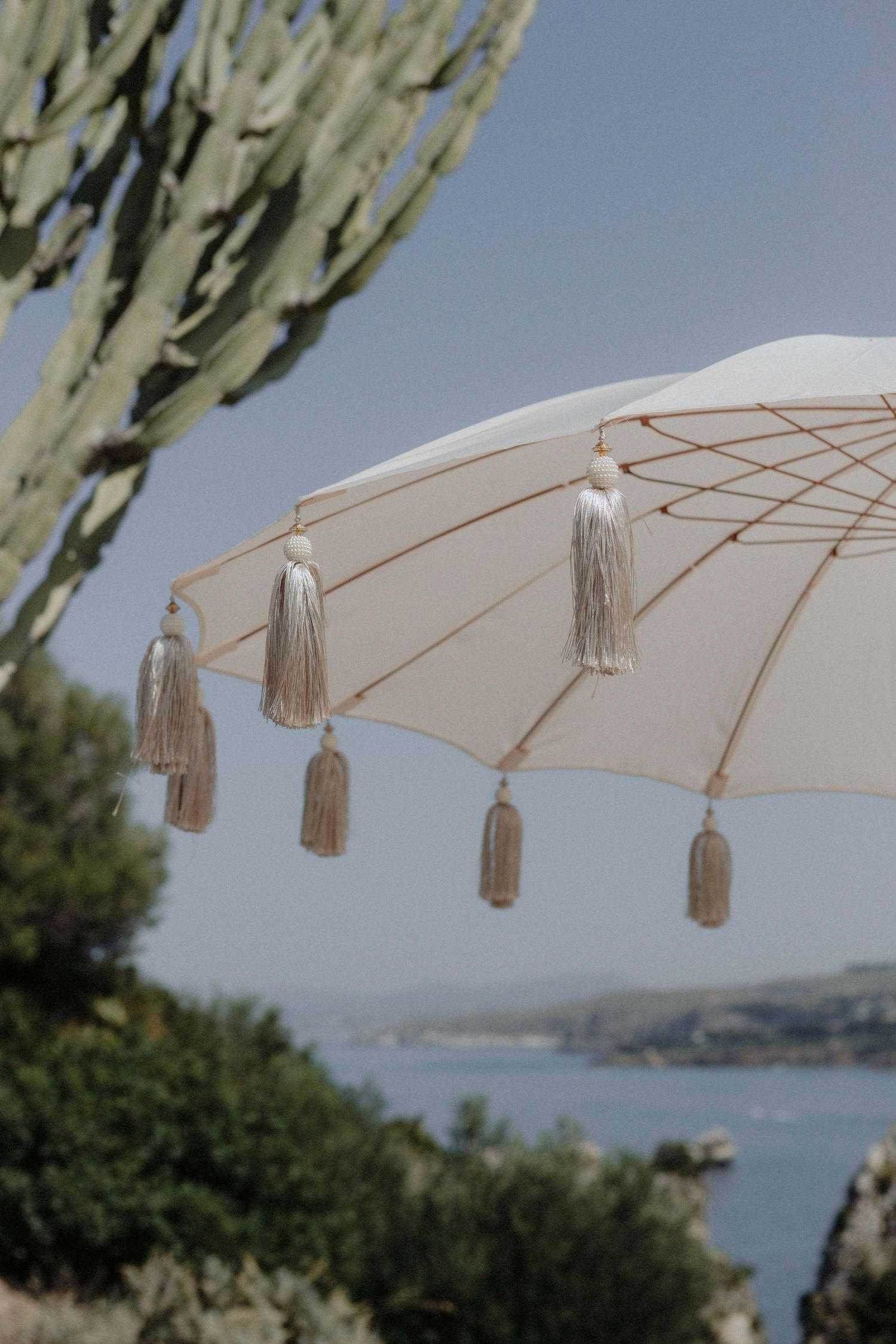 Elegant white parasol with tassels overlooks a coastal Mediterranean landscape with cacti and blue sea.