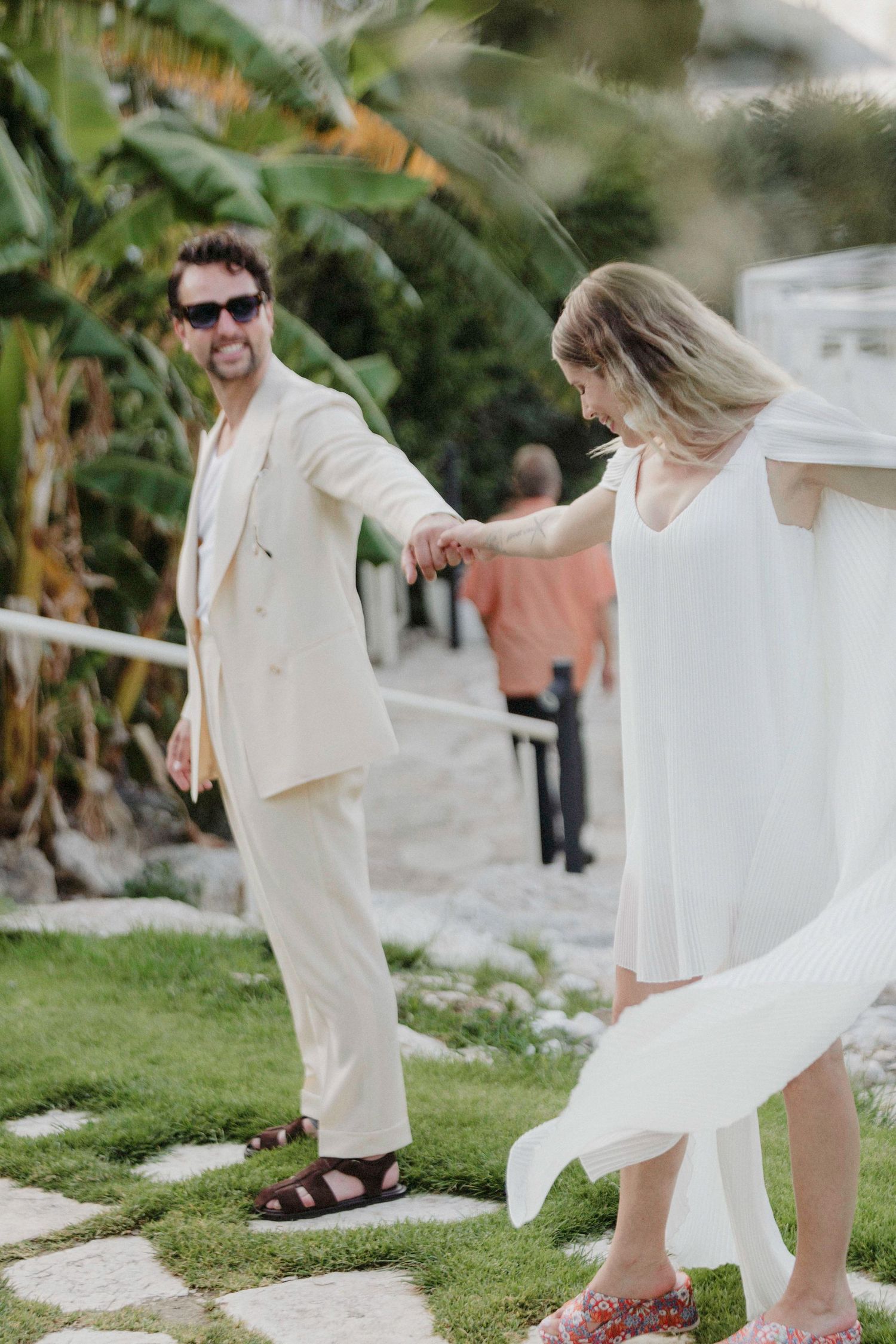 A smiling man in a cream suit holds hands with a woman in a white dress outdoors amid lush tropical greenery.