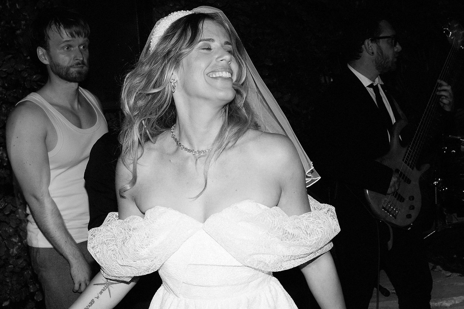 A bride in a strapless white wedding dress with puffy sleeves laughs joyfully at a reception.