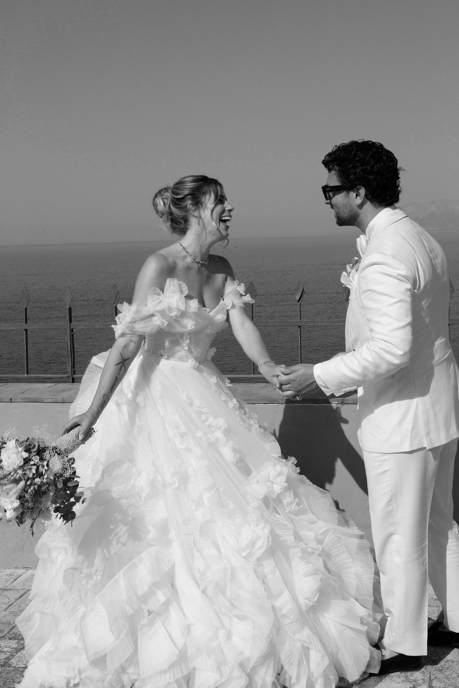 Black and white photo of a laughing bride in ruffled gown holding bouquet with groom in white suit by the sea.
