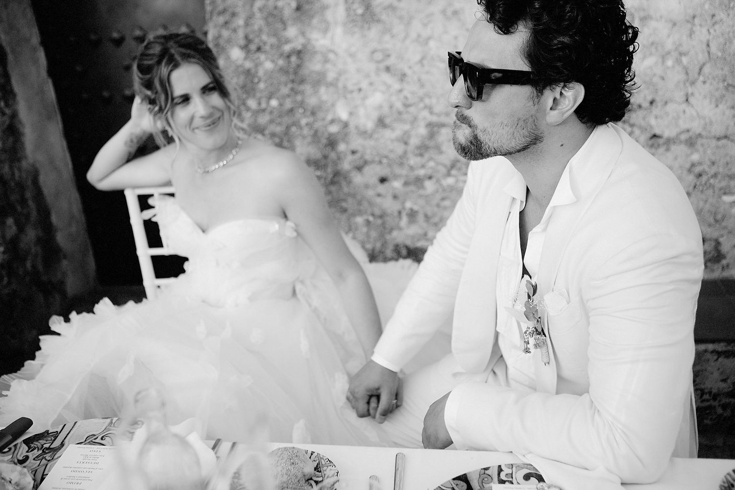 Black and white photo of a bride in a strapless gown and a groom in sunglasses seated at a table.
