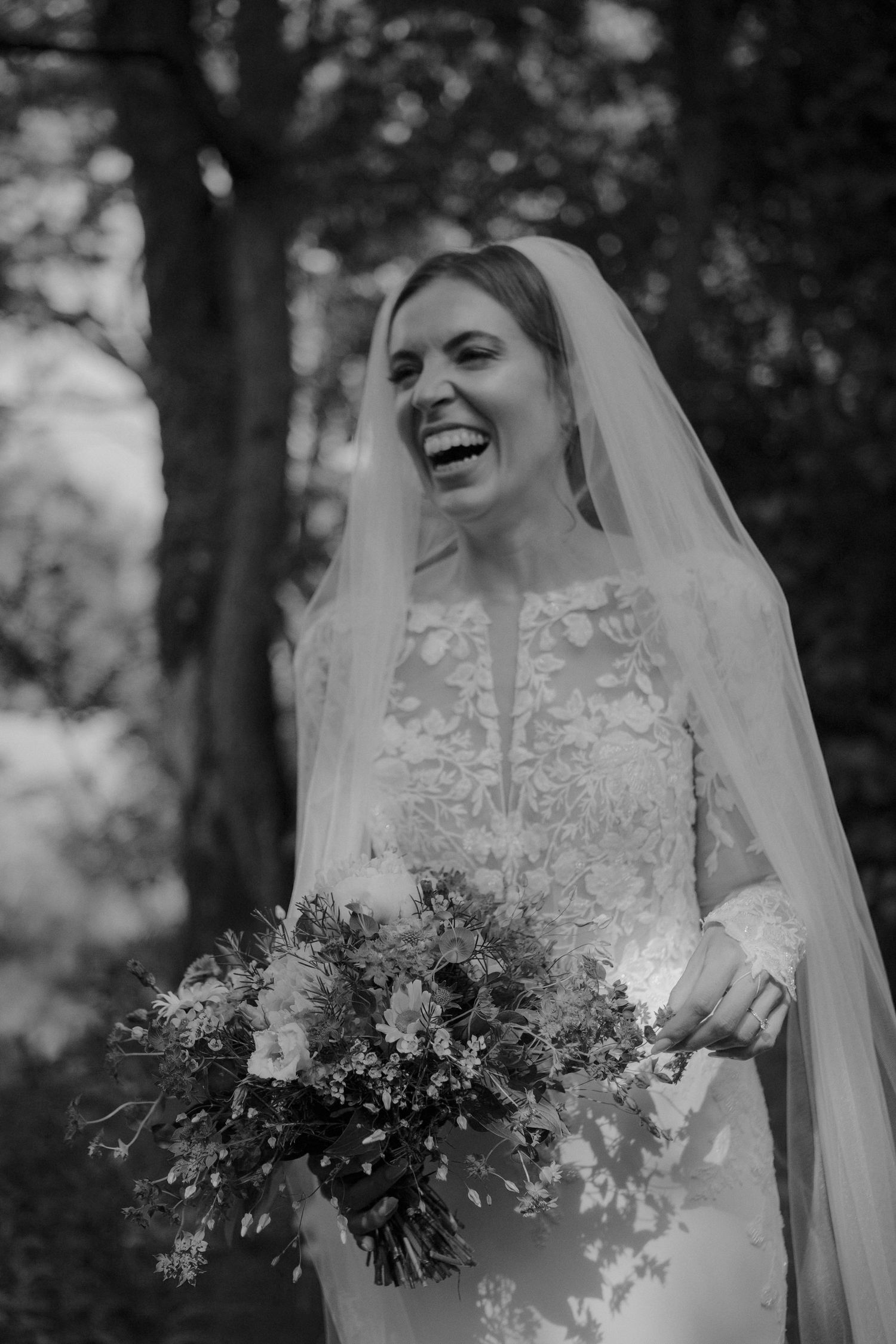A joyful bride in a lace wedding dress laughs while holding a wild flower bouquet in a black and white photo.