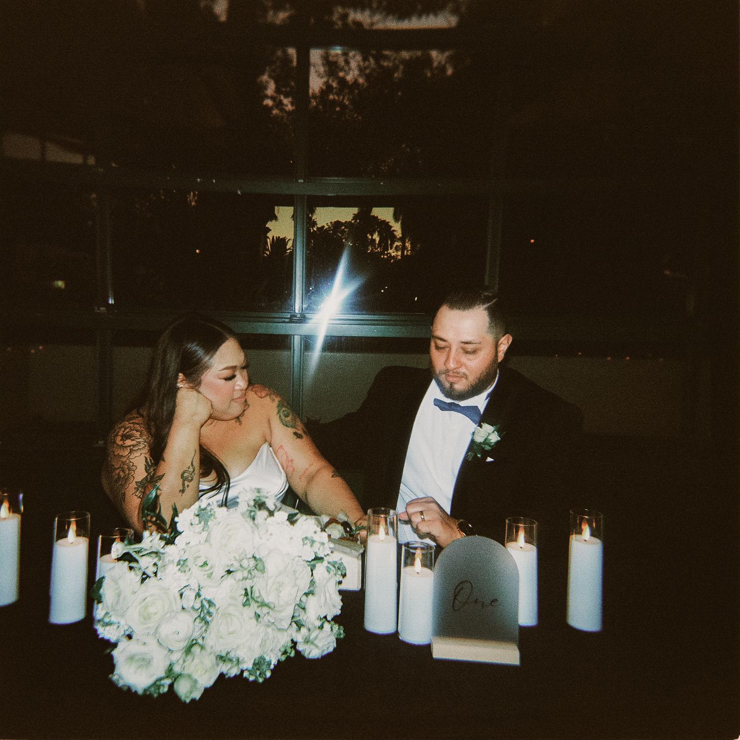 Couple sits at candlelit wedding reception table with white floral centerpiece and glowing pillar candles.