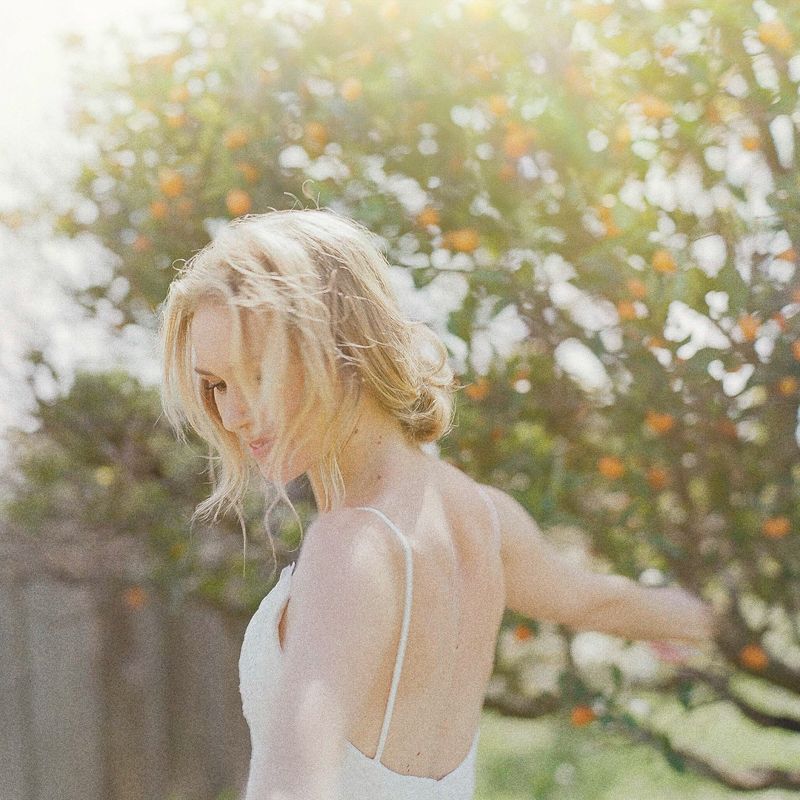 Blonde woman in white dress twirling outdoors near orange fruit tree in soft golden sunlight.