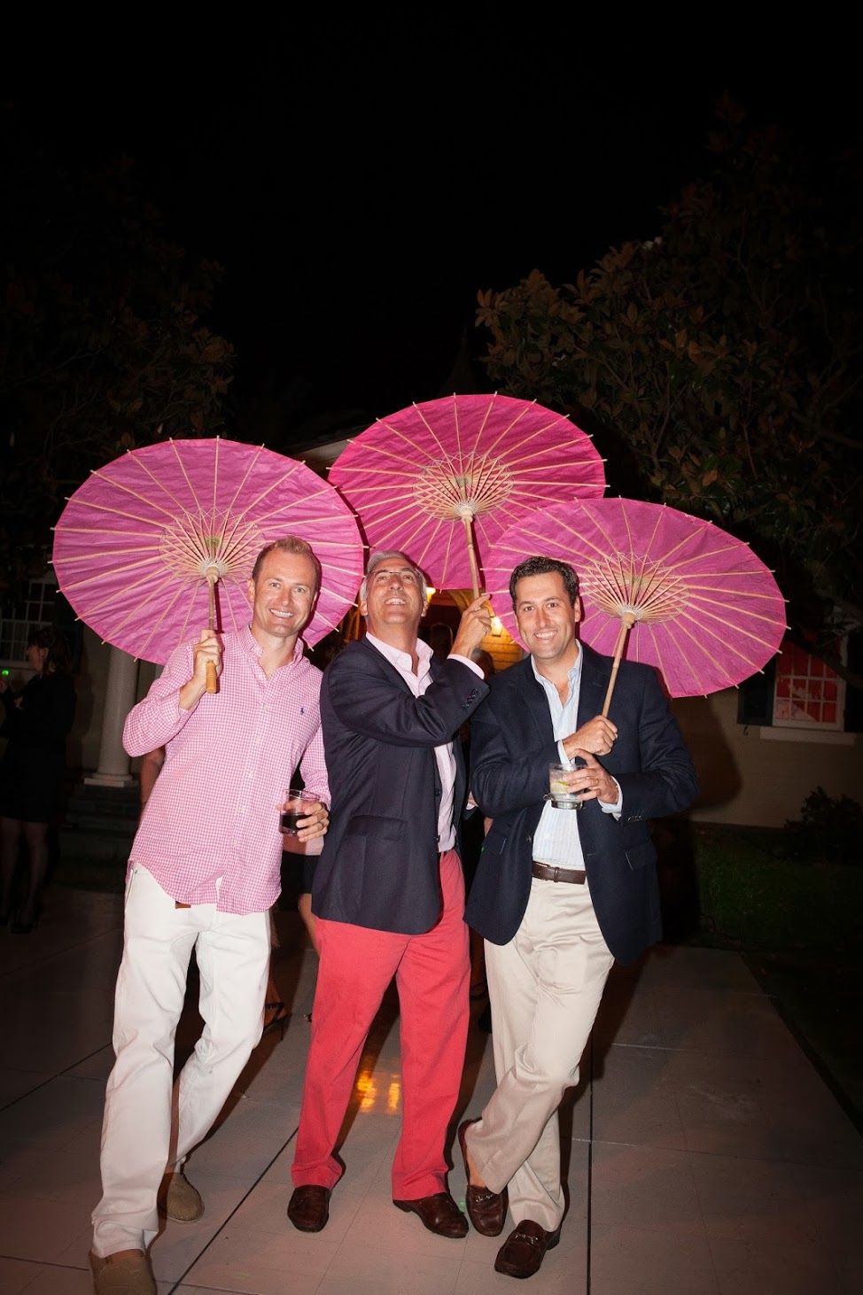 Three people in evening attire pose together holding pink paper parasols at a nighttime event.