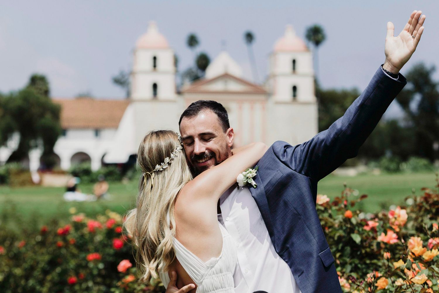 A joyful wedding celebration scene in a rose garden with a white church building in the distance.