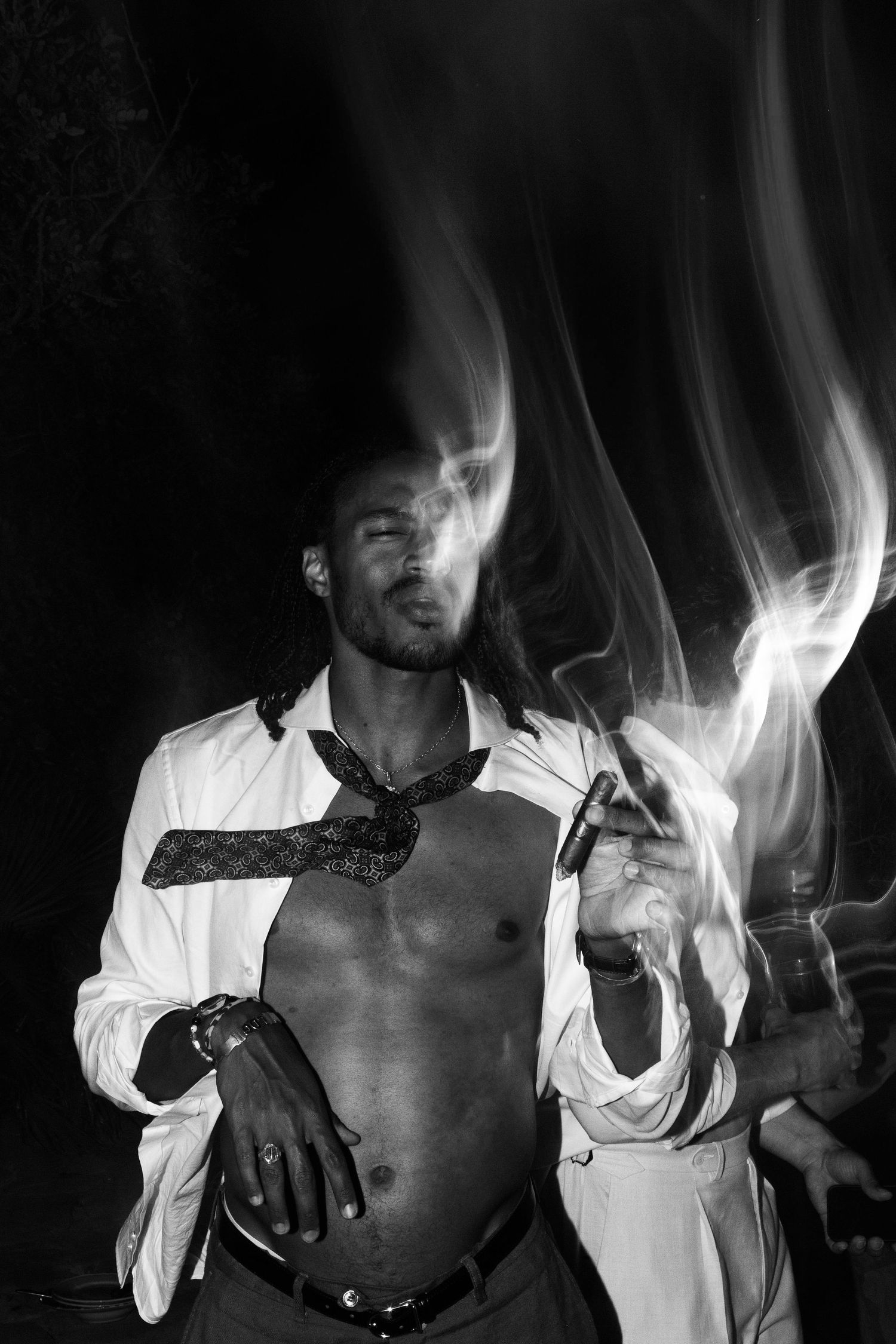 Black and white photo of a shirtless man in an open white shirt smoking a cigar with dramatic smoke swirls.