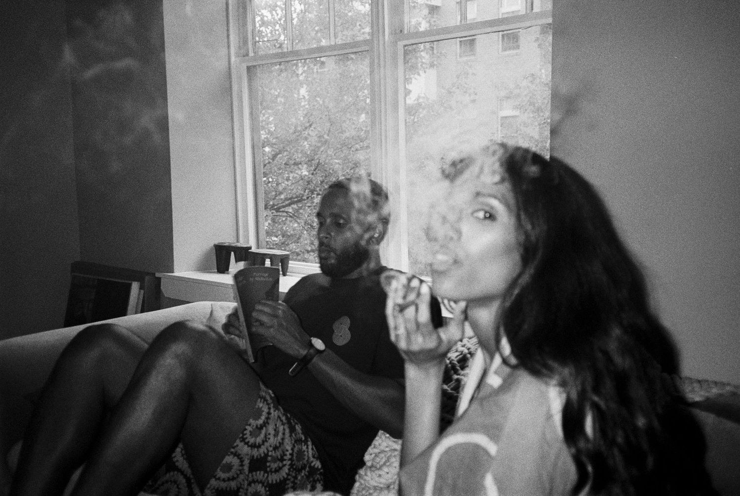 Black and white photo of two people relaxing indoors, with a woman blowing smoke near a window.