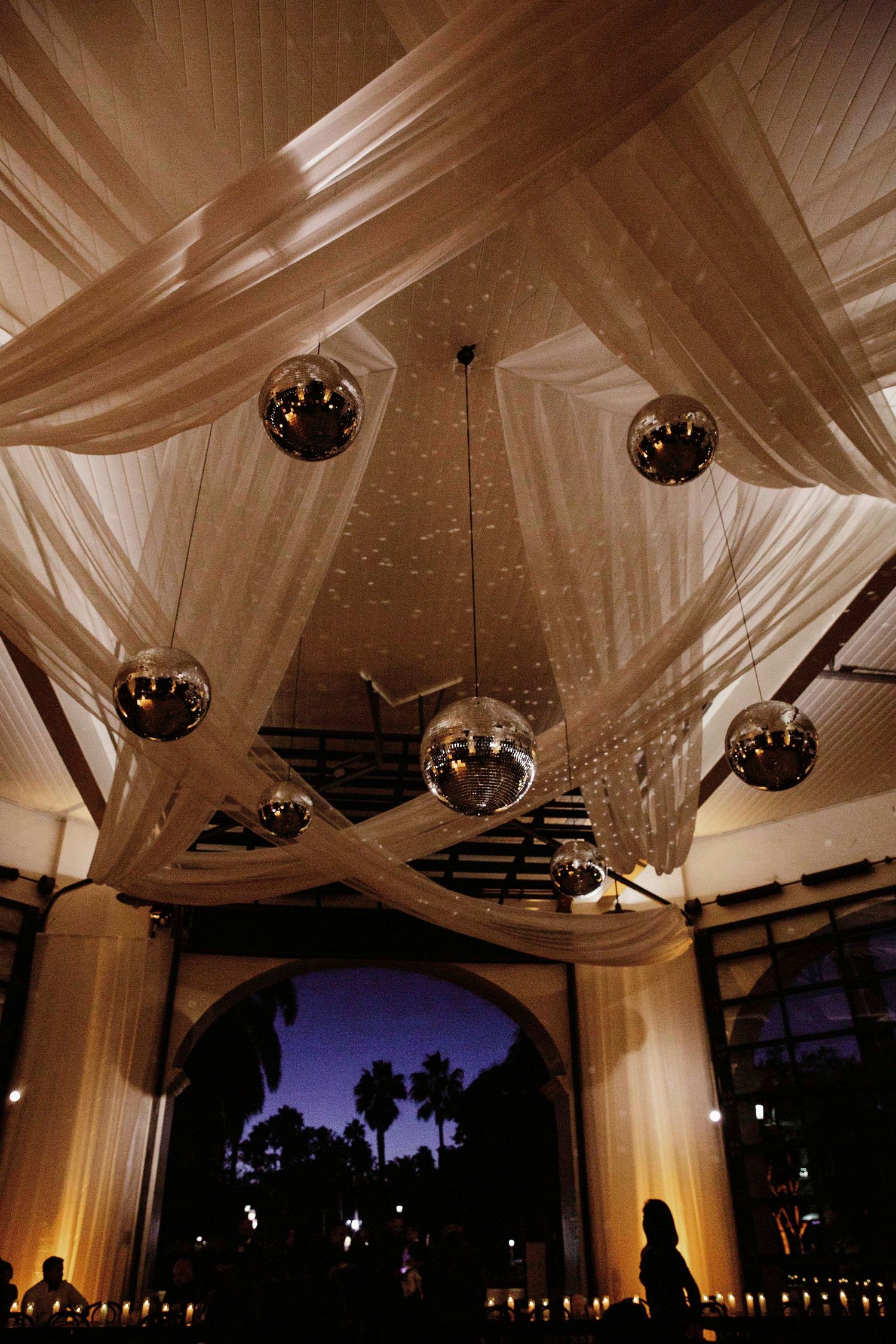 Elegant wedding reception tent with white draped fabric ceiling, disco balls, and palm trees silhouetted against sunset sky.