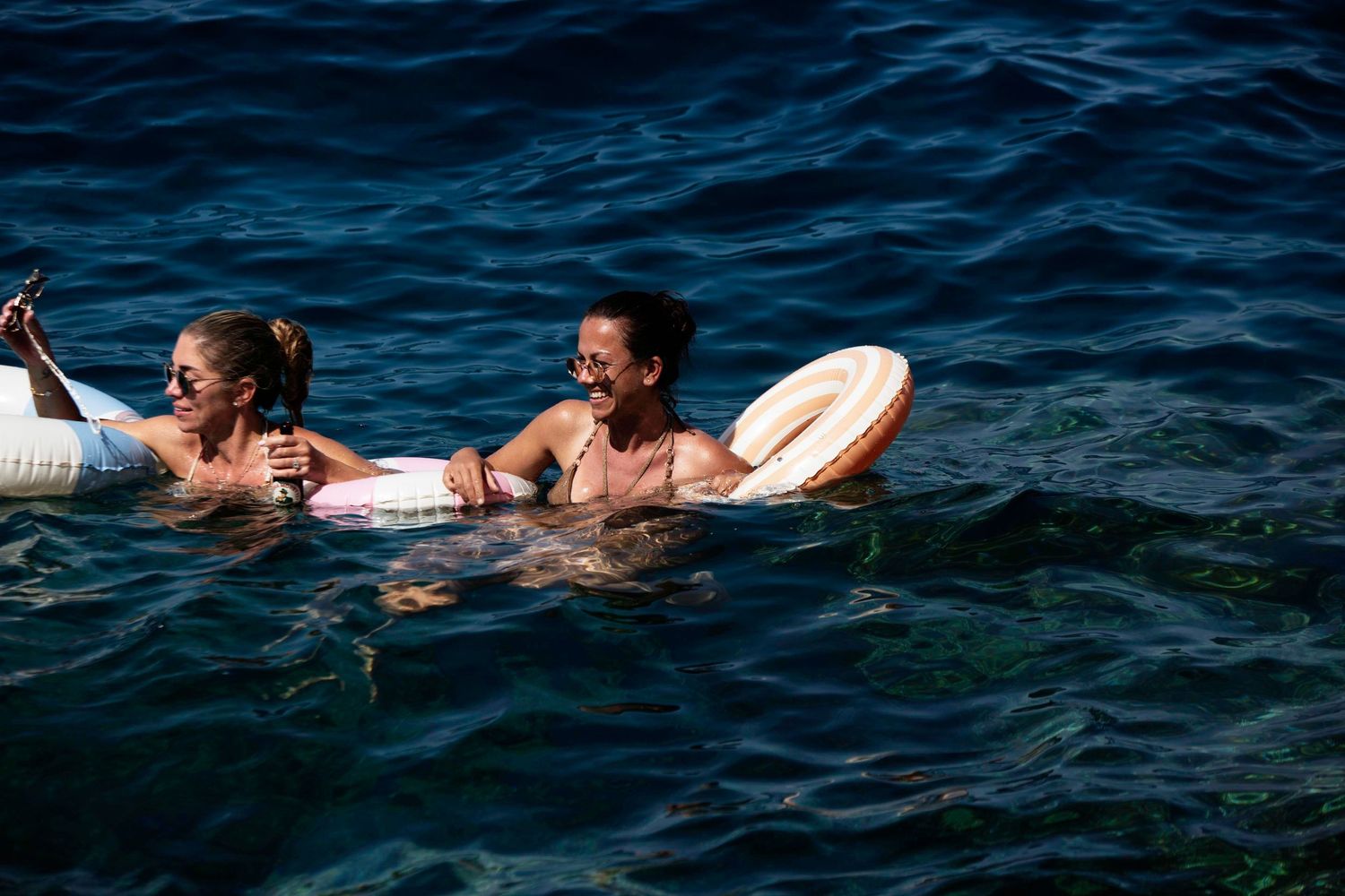 Two women swimming in clear blue ocean water while holding onto a white surfboard on a sunny day.
