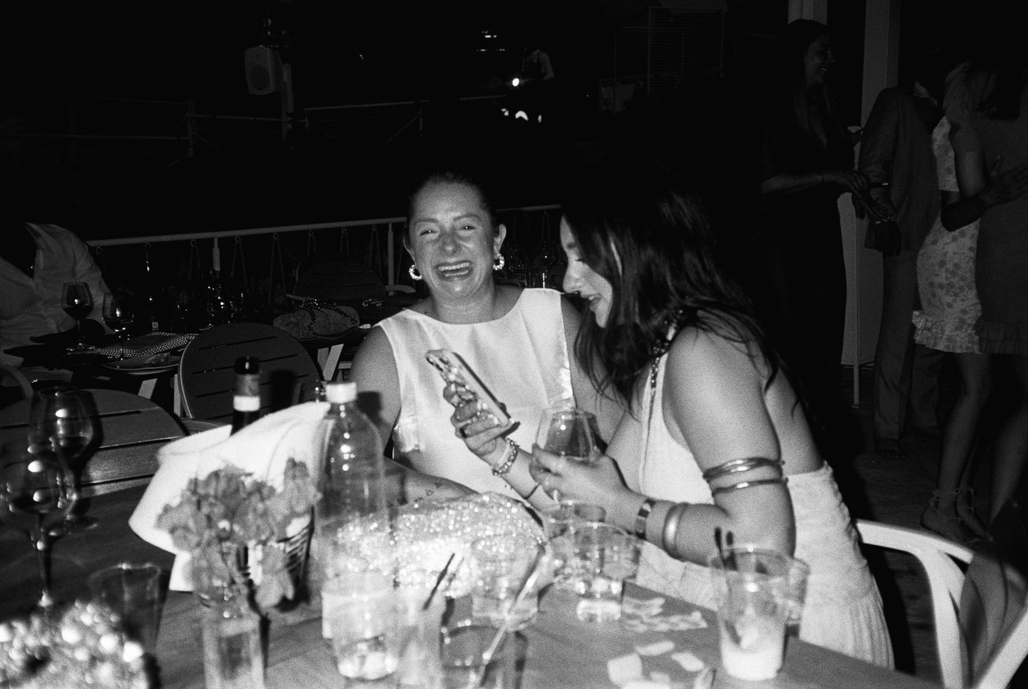 Two people share a candid moment at a table with drinks during what appears to be a nighttime social gathering.